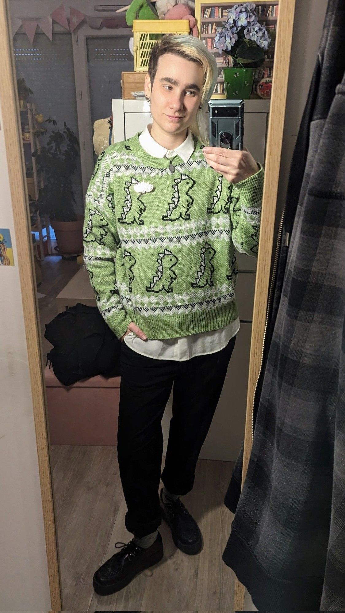 Selfie miroir dans mon appart. J'ai une main dans la poche et je regarde mon portable avec un léger sourire. Je porte une chemise blanche qui dépasse au col et en bas de mon pull vert tendre à motifs dinosaures. Je porte également un pantalon de costume noir retroussé, des chaussettes vert tendre et des creepers noires.