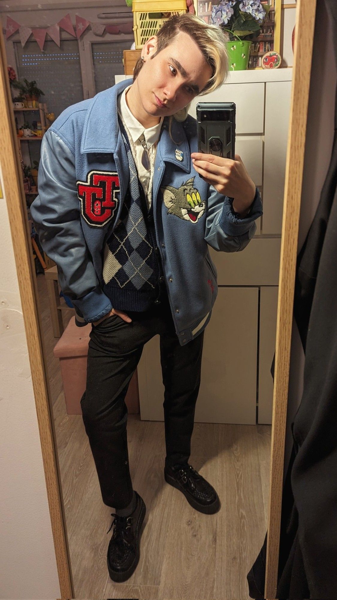 Selfie miroir dans mon appart. J'ai une main dans ma poche de pantalon, je me penche légèrement en avant et je regarde mon portable avec un léger sourire. Je porte un blouson Teddy bleu avec un patch rouge et un patch Tom de Tom et Jerry dessus. Je porte également une chemise en dentelle blanc cassé avec un cardigan assez court à motifs jacquard bleu et gris, un pendentif aile de papillon bleu en guise de cravate, un pantalon de costume gris foncé, des chaussettes gris foncé et des creepers noires.