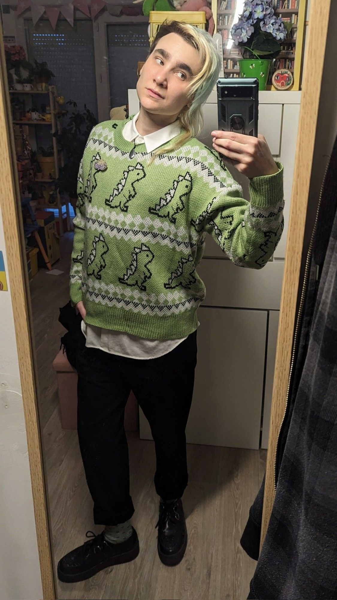 Selfie miroir dans mon appart. Je suis de 3/4 face du côté des cheveux longs, j'ai une main dans la poche et je regarde mon portab. Je porte une chemise blanche qui dépasse au col et en bas de mon pull vert tendre à motifs dinosaures. Je porte également un pantalon de costume noir retroussé, des chaussettes vert tendre et des creepers noires.