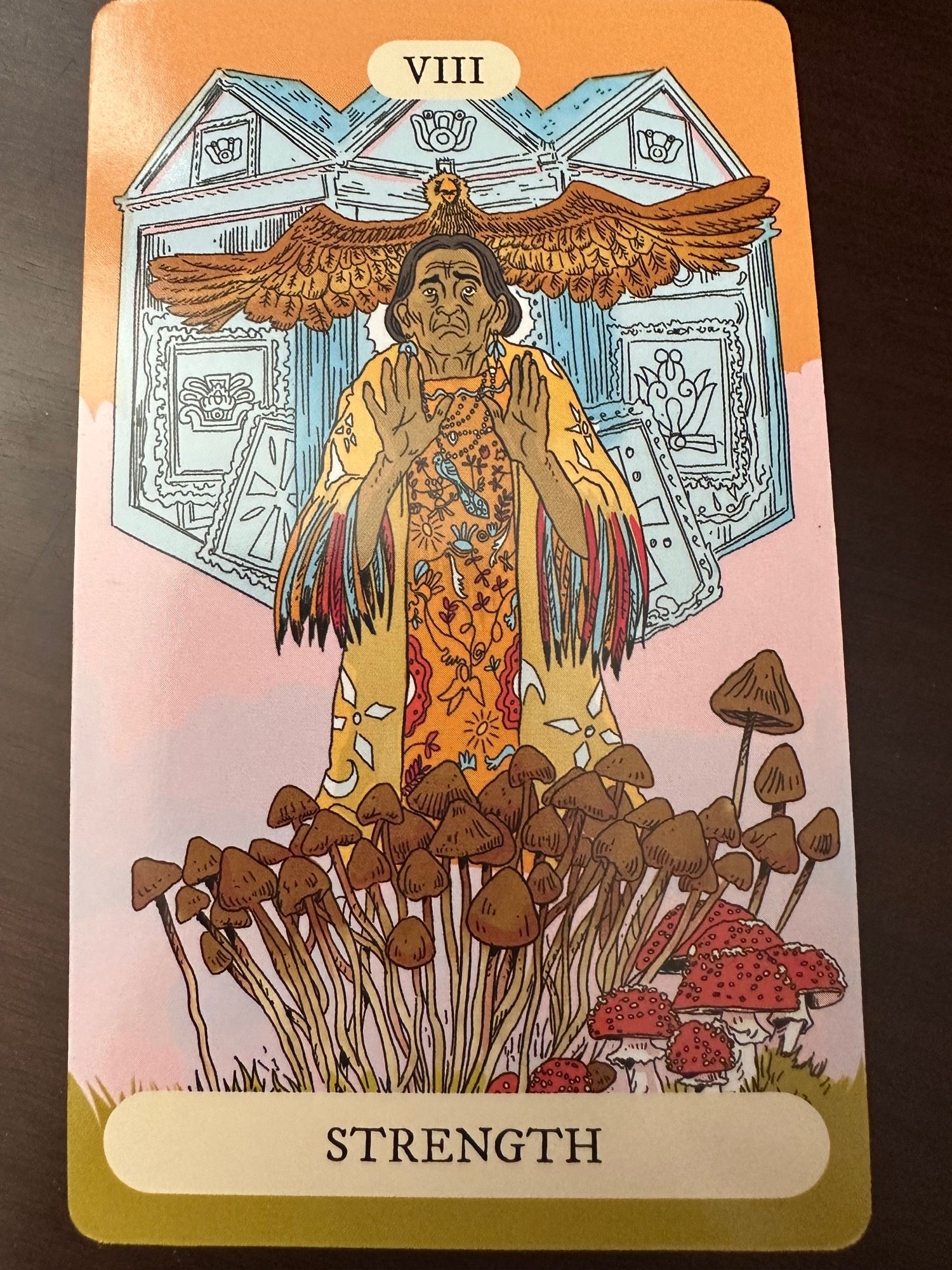 Photo of the 8 cars from my tarot deck, Strength. María Sabina is the featured character for this card, she’s wearing what looks like traditional Mexican healer clothing behind a lot of tall brown mushrooms and a few short red and white mushrooms next to them. Behind her is an eagle and what looks to be a building related to healing.