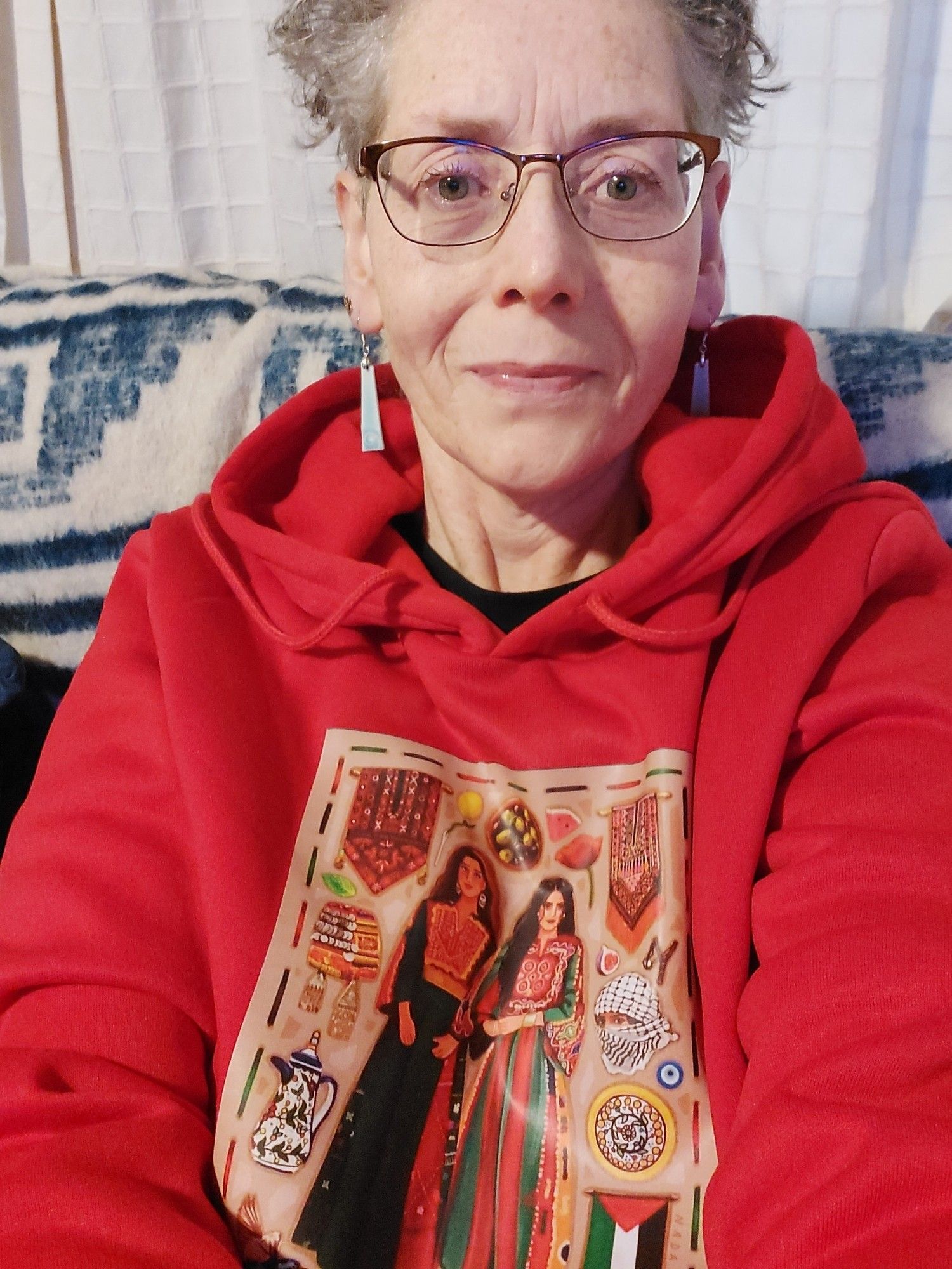 A selfie of me, a pākeha woman with silver hair & glasses in a red hoodie with print of 2 Palestinian women & various cultural taonga.