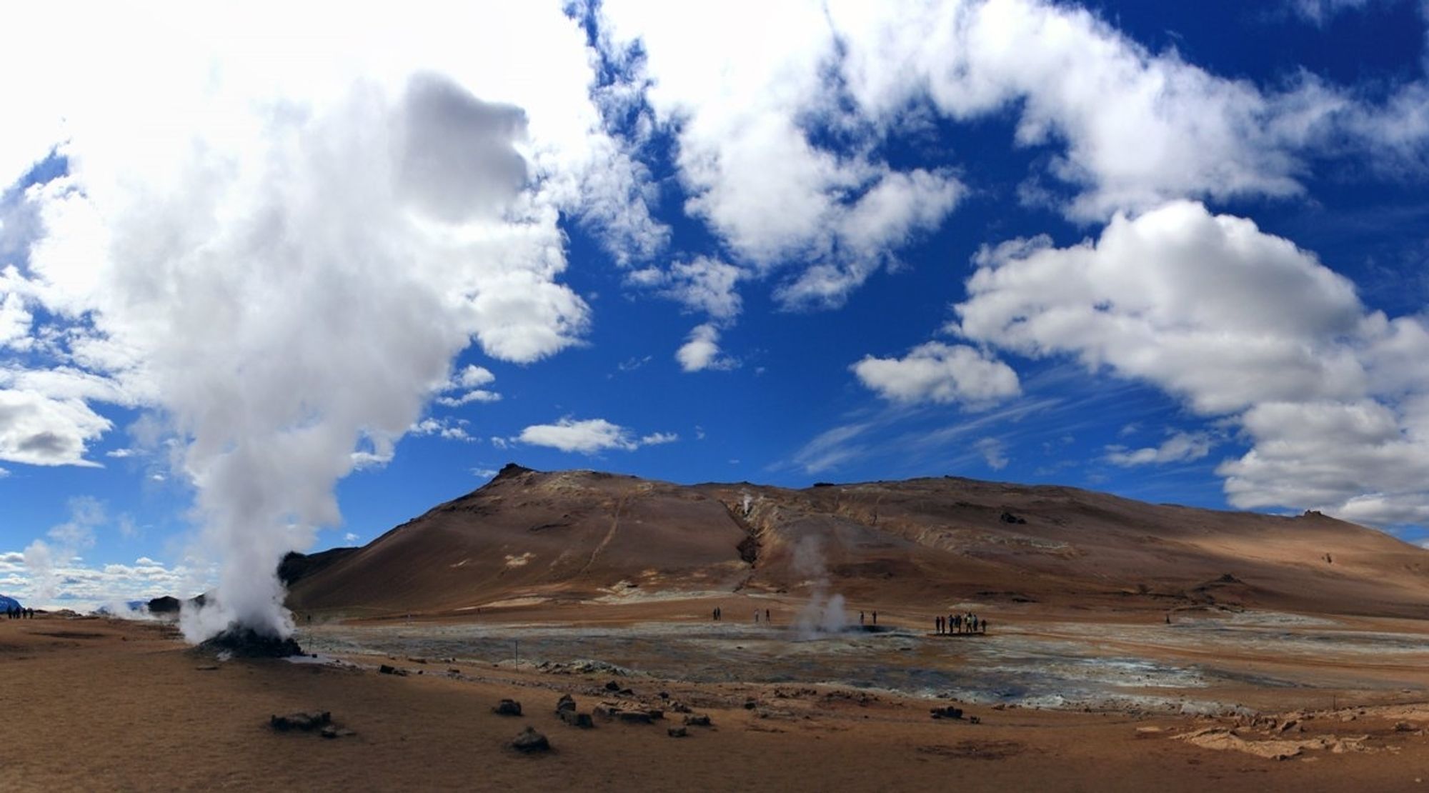 Foto in Námafjall, ein Bergrücken in Island im Mývatn-Gebiet. Die Landschaft ist rot-braun mit vereinzelten grauen Flächen. Durch die vulkanische Aktivität steigt am linken Bildrand weißer Rauch auf. Auch in der Bildmitte ist über einem kleinen Krater grauer Rauch zu sehen. Der Himmel ist kräftig blau mit weißen Wolken