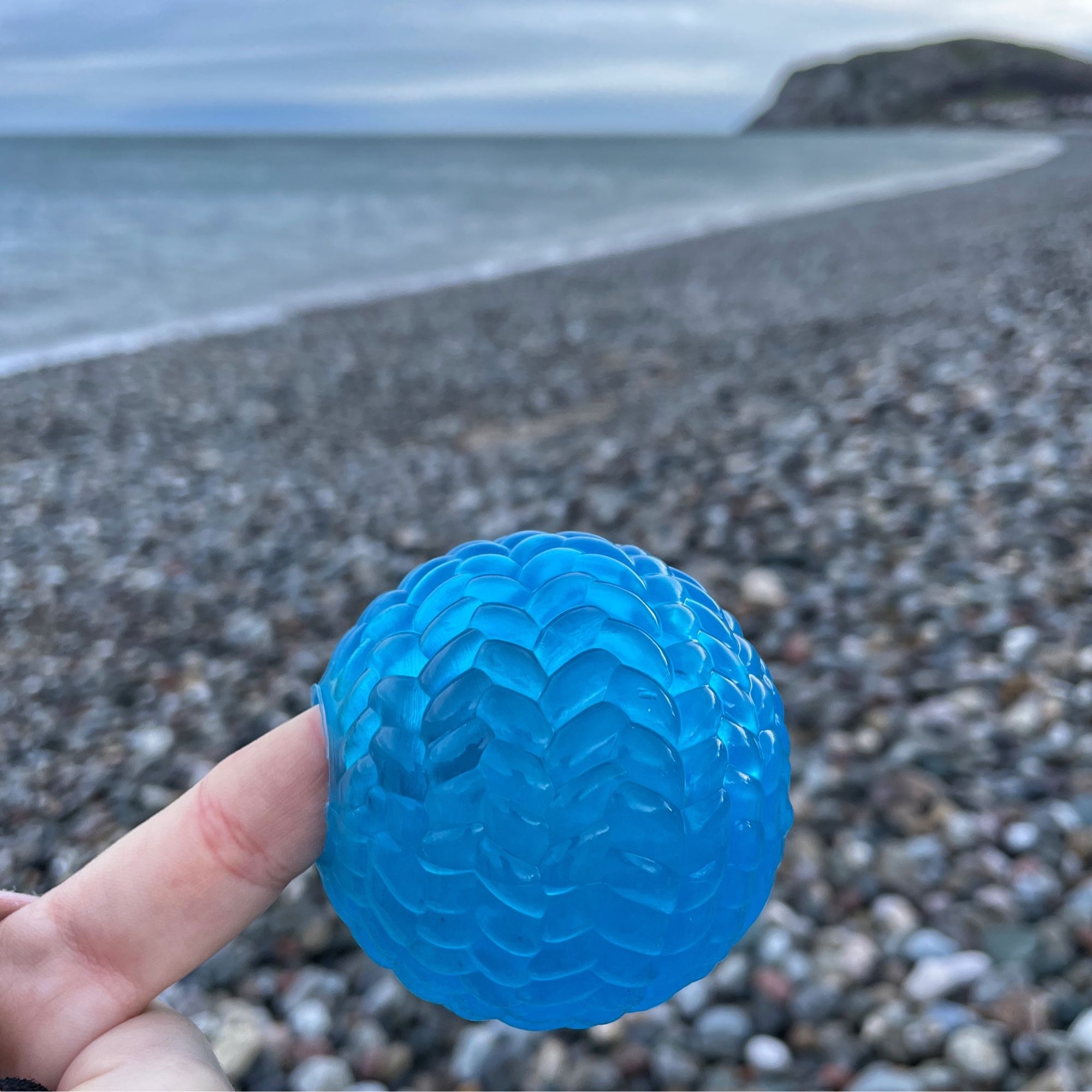 The same blue plastic ball held up by a finger to show scale (approx 8-10cm diameter) background is a pebbly beach.