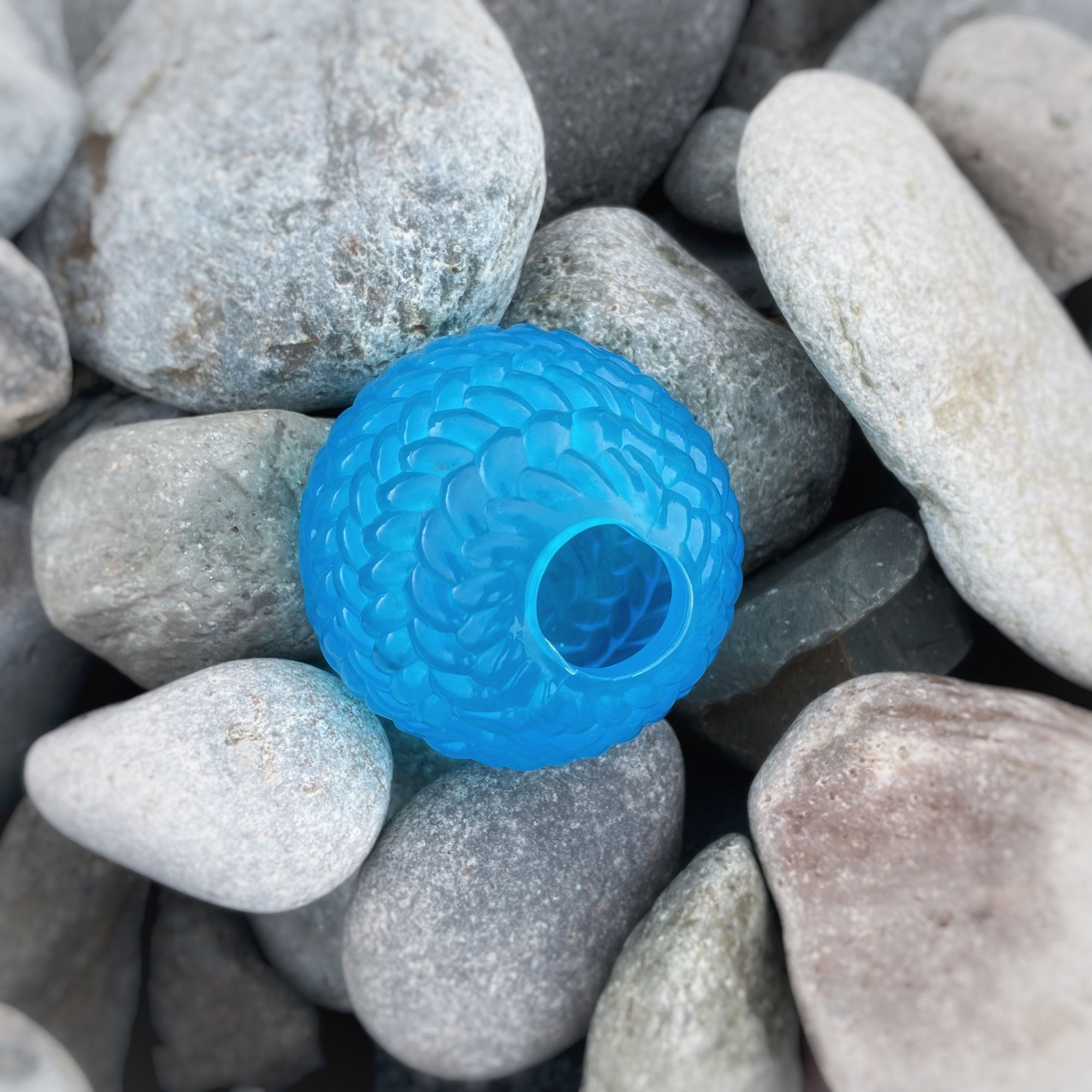 A bright blue plastic hollow ball with a textured surface resting on smooth pale grey stones