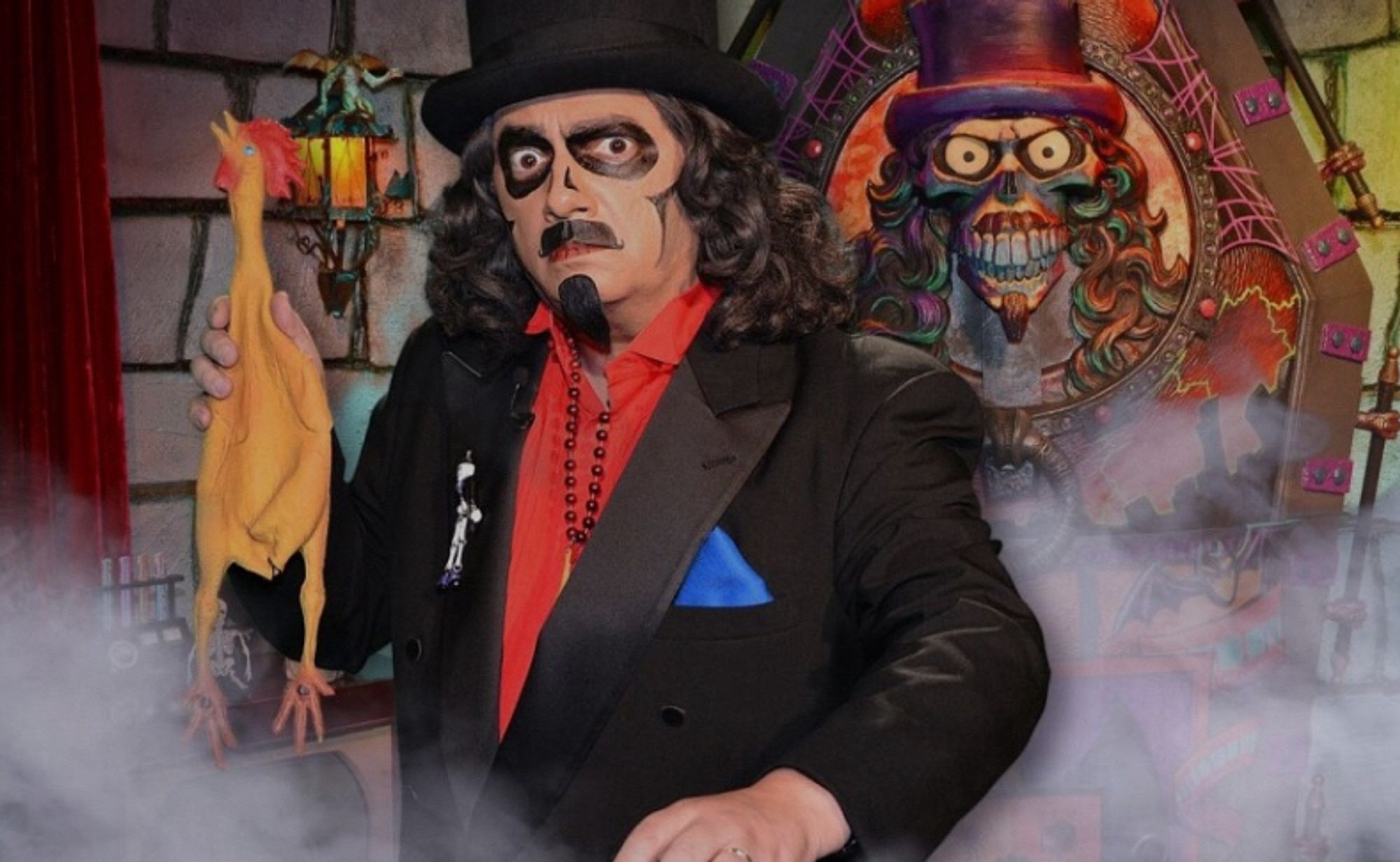 Horror host Svengoolie stares into camera with rubber chicken named Kerwyn, in his dungeon lair with fog and his heavily decorated casket behind him