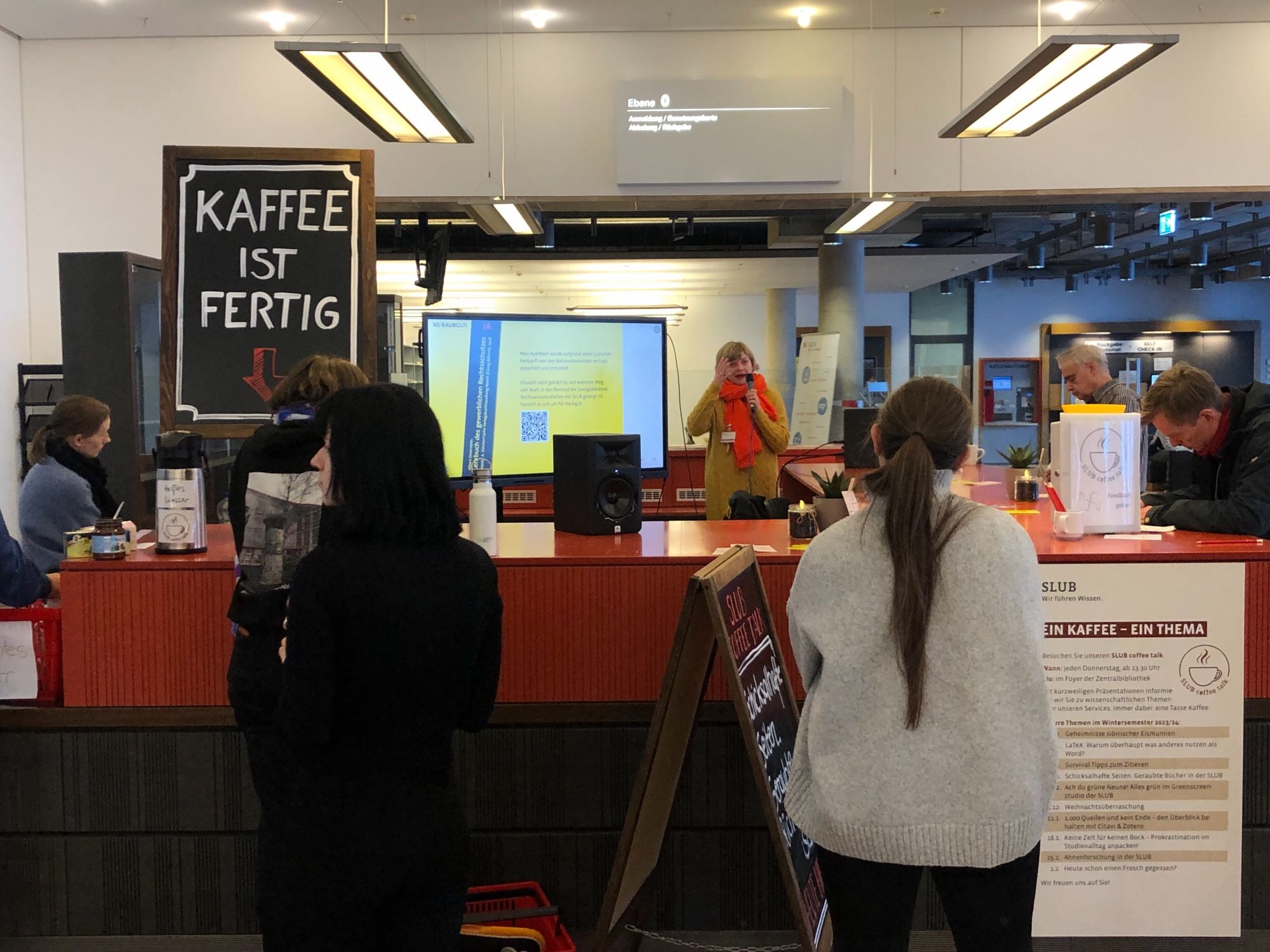 Zu sehen ist eine Personengruppe. Es spricht eine Frau mit einem gelben Pullover.
Im linken Bildrand sieht man ein Schild mit dem Schriftzug „Kaffee ist fertig“.