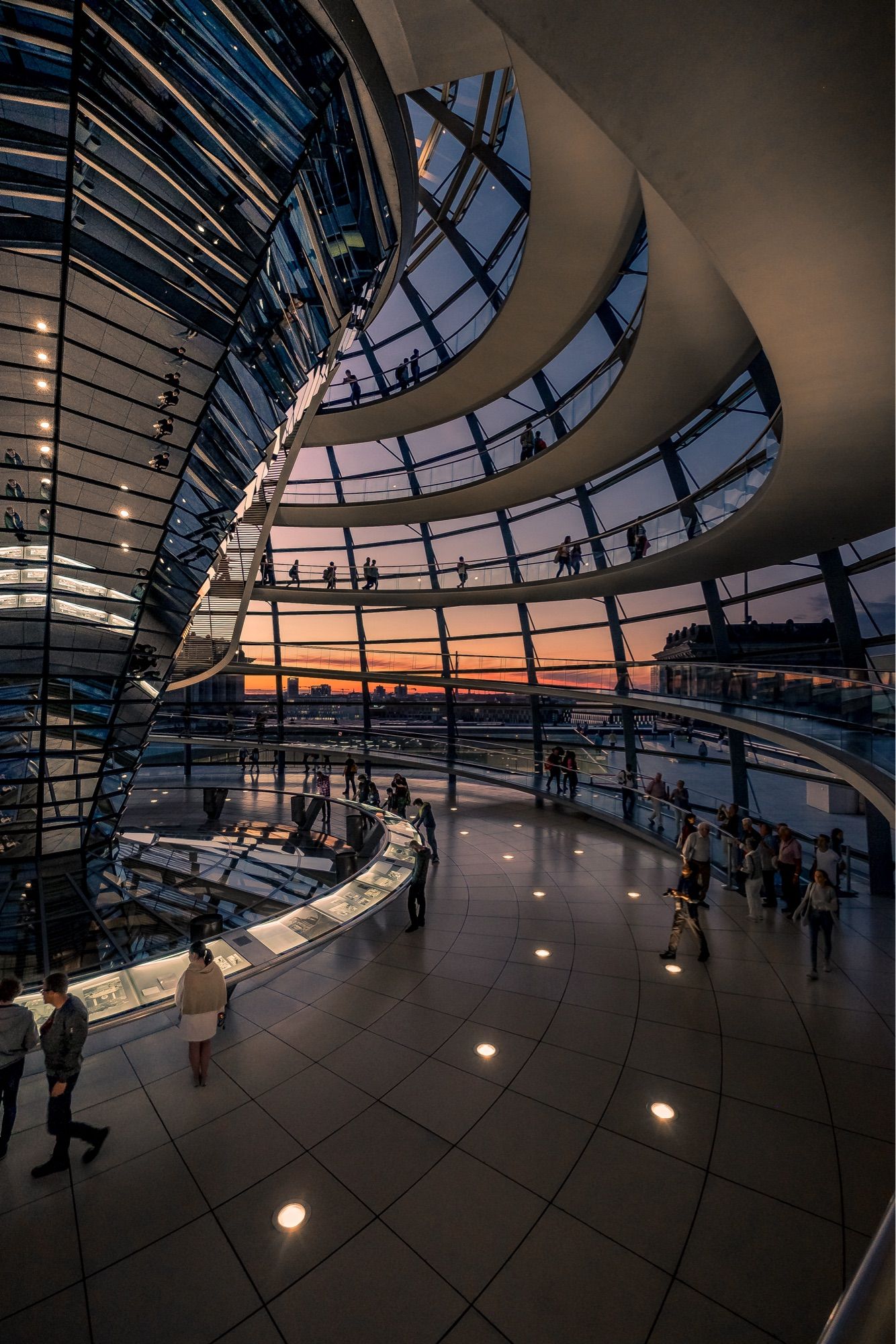 Das Bild zeigt die beeindruckende Innenseite der Kuppel des Reichstagsgebäudes in Berlin. Die Kuppel ist aus Glas und Stahl und bietet einen atemberaubenden Blick auf die Stadt. 

Im Vordergrund sind mehrere Besucher zu sehen, die die exponierte Struktur und die Ausstellungen bewundern. Die spiralförmige Treppe führt nach oben, während die Wände mit reflektierenden Oberflächen versehen sind, die das Licht einfangen und verstärken. 

Der Sonnenuntergang im Hintergrund sorgt für eine warme, goldene Beleuchtung, die die Atmosphäre des Raumes zusätzlich verstärkt. Die Kombination aus modernem Design und historischer Bedeutung macht diesen Ort einzigartig und ein beliebtes Ziel für Touristen.