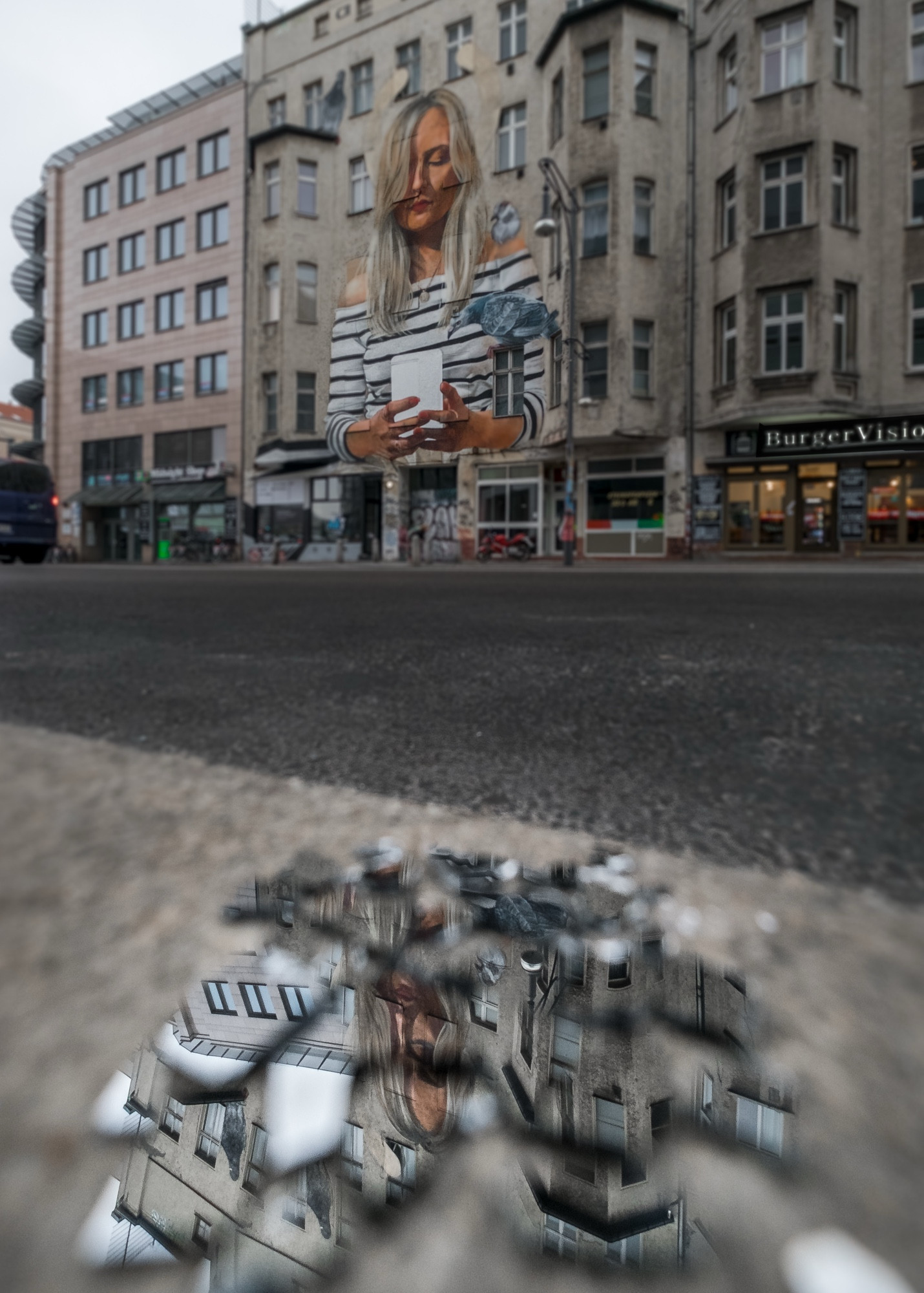 Das Bild zeigt eine Straßenszene in einer urbanen Umgebung, vermutlich in einer deutschen Stadt, erkennbar an den architektonischen Details und den Schildern im Hintergrund. Der Fokus des Bildes liegt auf einem beeindruckenden Wandgemälde, das an der Fassade eines Gebäudes zu sehen ist. Das Gemälde zeigt eine blonde Frau, die auf ihr Handy schaut. Sie trägt ein gestreiftes Oberteil und ist sehr detailliert dargestellt, fast wie ein realistisches Porträt, das sich nahtlos in die Gebäudeoberfläche einfügt.

Im Vordergrund spiegelt sich ein Teil dieses Wandgemäldes in Spiegelscherben, die auf dem Boden liegen. Diese Scherben fügen der Komposition eine interessante und ungewöhnliche visuelle Ebene hinzu. Der nasse oder raue Asphalt, der leicht unscharf ist, sorgt für Tiefe und Struktur im Bild. Die Reflexion in den Scherben bietet eine clevere Perspektive und zieht den Blick des Betrachters auf diese unerwartete Darstellung des Gemäldes, die aus einem anderen Winkel erscheint.

Die umliegenden Gebäude und Geschäfte, wie „BurgerVision“, verstärken das städtische Setting und geben dem Bild eine alltägliche, aber zugleich künstlerische Note. Die Gesamtkomposition spielt gekonnt mit Schärfe, Unschärfe und Reflexionen in den Spiegelscherben, was dem Bild eine besondere Dynamik und Originalität verleiht.