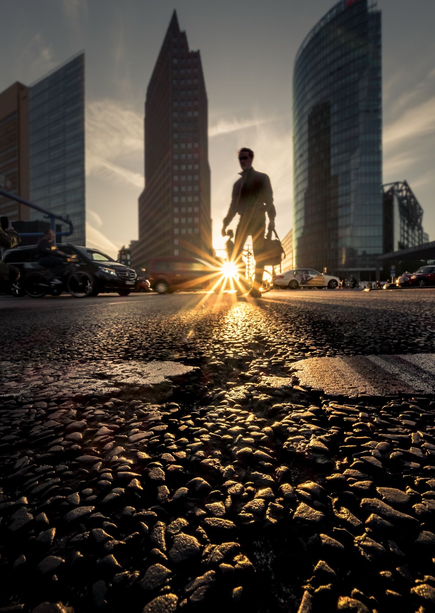 Das Bild zeigt eine städtische Szene am Potsdamer Platz in Berlin. Ein Mann in einem Anzug geht auf der Straße und hält eine Tasche in der Hand. Die Sonne scheint hinter ihm und erzeugt einen strahlenden Effekt, der die Szene beleuchtet. Im Hintergrund sind moderne Gebäude zu sehen, die den urbanen Charakter der Umgebung unterstreichen. Im Vordergrund sieht man den Asphalt der Straße, der durch das Licht und die Perspektive wie kleine Steine wirkt, was der Atmosphäre eine besondere Note verleiht.