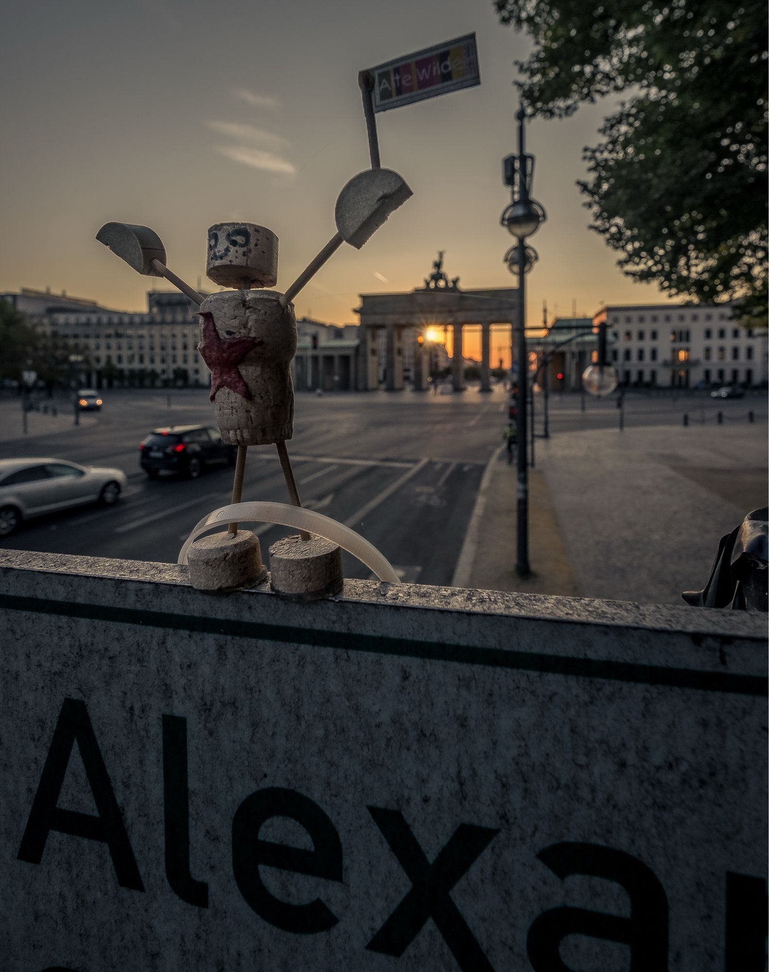 Das Bild zeigt eine kleine Figur, die aus Weinkorken und anderen recycelten Materialien zusammengebastelt wurde. Die Figur hat einen bemalten Kopf und hält zwei Schilder in den Händen. Auf einem Schild steht “Alte Wilde.” Die Figur steht auf einem Straßenschild, das teilweise zu erkennen ist und wahrscheinlich den Namen “Alexanderplatz” zeigt.

Im Hintergrund ist das Brandenburger Tor in Berlin zu sehen, das von der untergehenden Sonne im warmen, goldenen Licht beleuchtet wird. Autos fahren auf der Straße, die zum Brandenburger Tor führt, während die Szenerie eine ruhige und atmosphärische Stimmung erzeugt, in der die warme Beleuchtung und der weite Himmel dominieren.

Die kreative und humorvolle Platzierung der Figur im Vordergrund kontrastiert mit der ikonischen Architektur des Brandenburger Tors im Hintergrund, was dem Bild eine spielerische, leicht surreale Note verleiht.