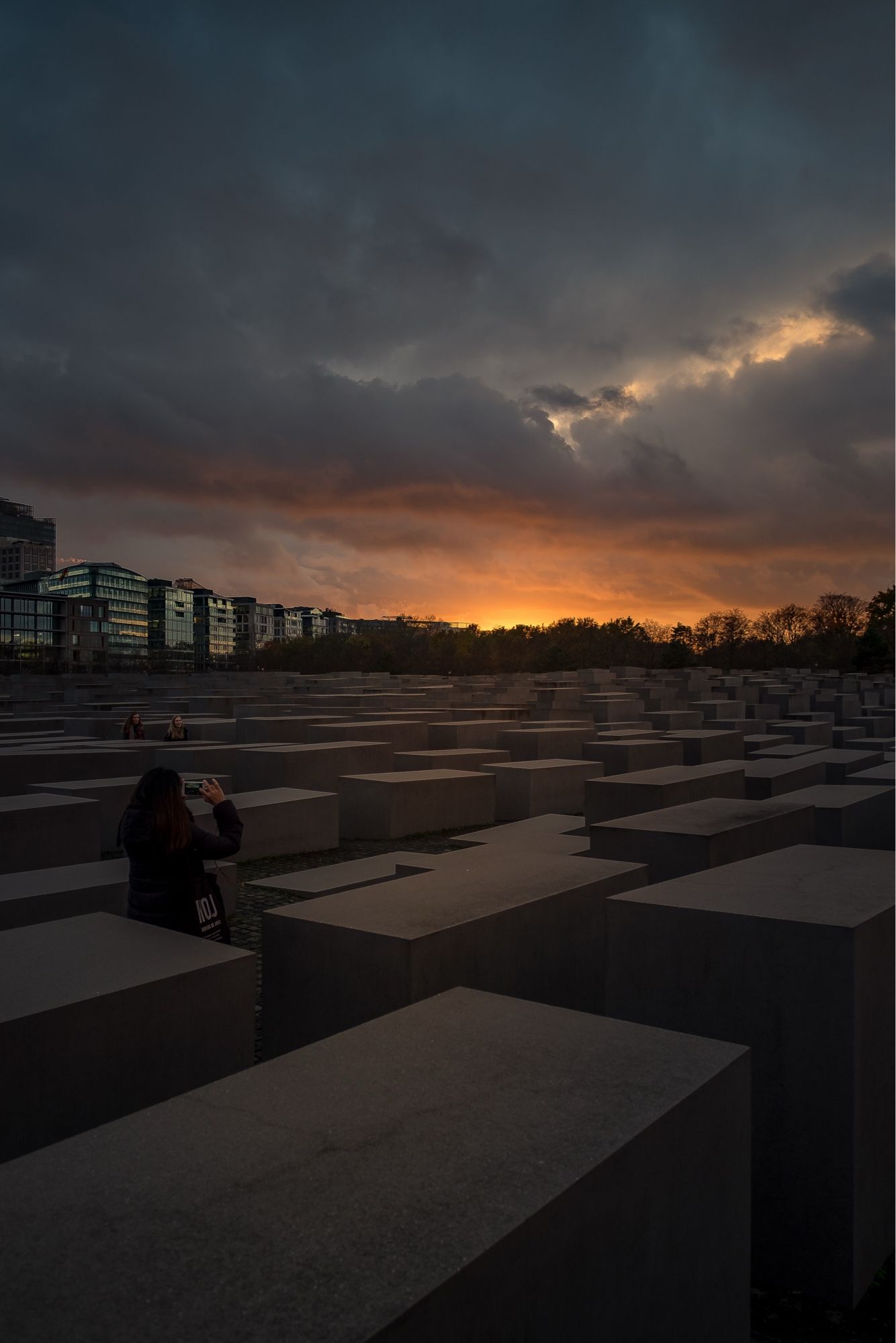 Das Bild zeigt das Holocaust-Mahnmal in Berlin, auch bekannt als das Denkmal für die ermordeten Juden Europas. Die Szene wurde während eines dramatischen Sonnenuntergangs aufgenommen, bei dem der Himmel von dunklen Wolken und einem intensiven orange-goldenen Licht dominiert wird, das sich am Horizont ausbreitet. Die Steinblöcke, die das Mahnmal ausmachen, erstrecken sich in einer geometrischen Anordnung, die aus vielen grauen, rechteckigen Betonstelen besteht.

Im Vordergrund sieht man eine Person, die das Mahnmal mit einem Handy fotografiert, während im Hintergrund weitere Besucher zwischen den Blöcken sichtbar sind. Die Gebäude der Stadt im Hintergrund fügen einen urbanen Kontrast zur Szenerie hinzu. Der düstere Himmel und das Licht der untergehenden Sonne verstärken die Atmosphäre des Ortes, der dazu dient, an die Opfer des Holocaust zu erinnern.