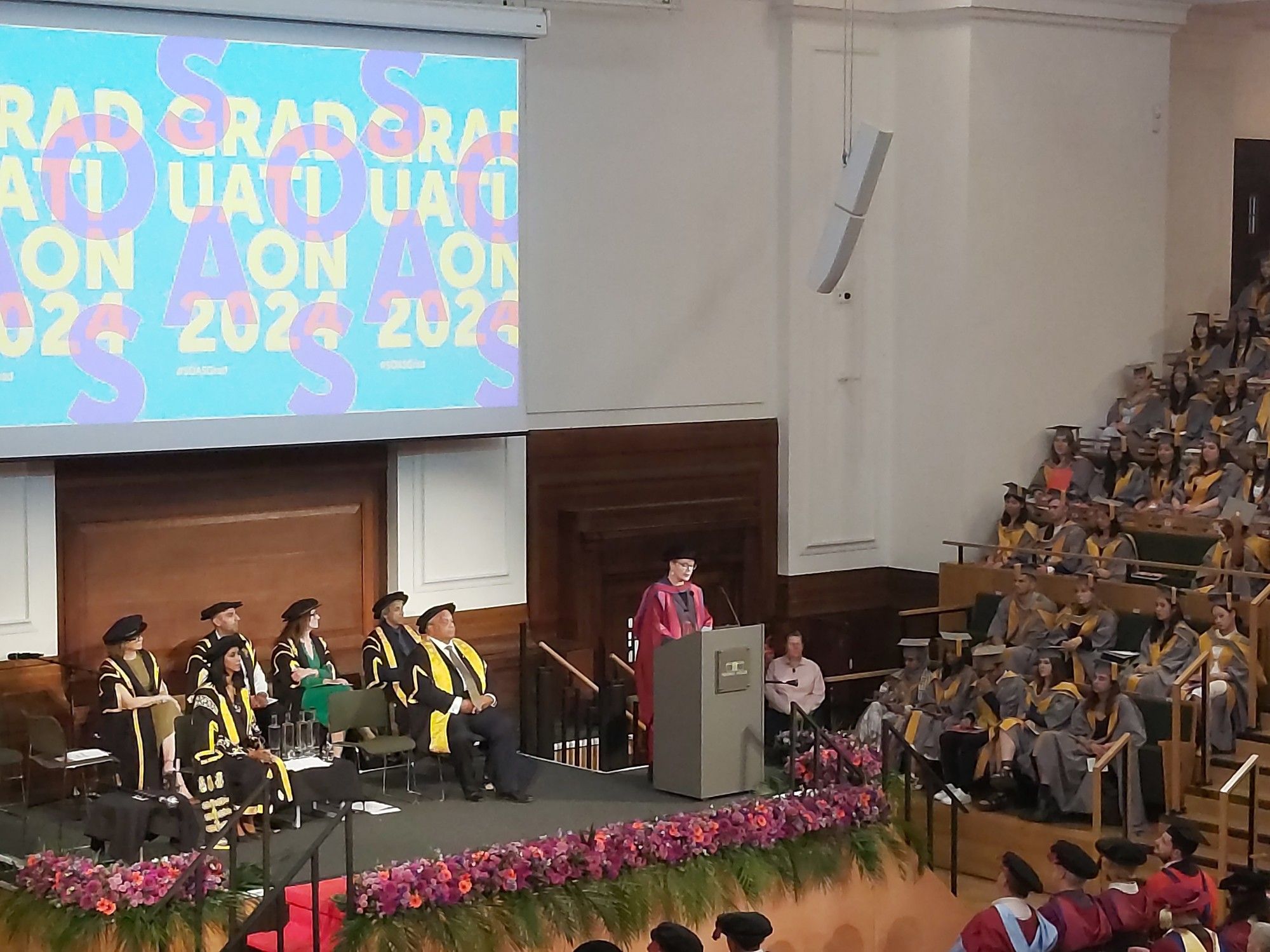 Graduation ceremony for SOAS University of London at Friends House, with robed faculty on the stage, and robed graduands on stepped seating. Prof. Rachel Harris is making the honorary citation for Rahile Dawut at the lectern on the stage.