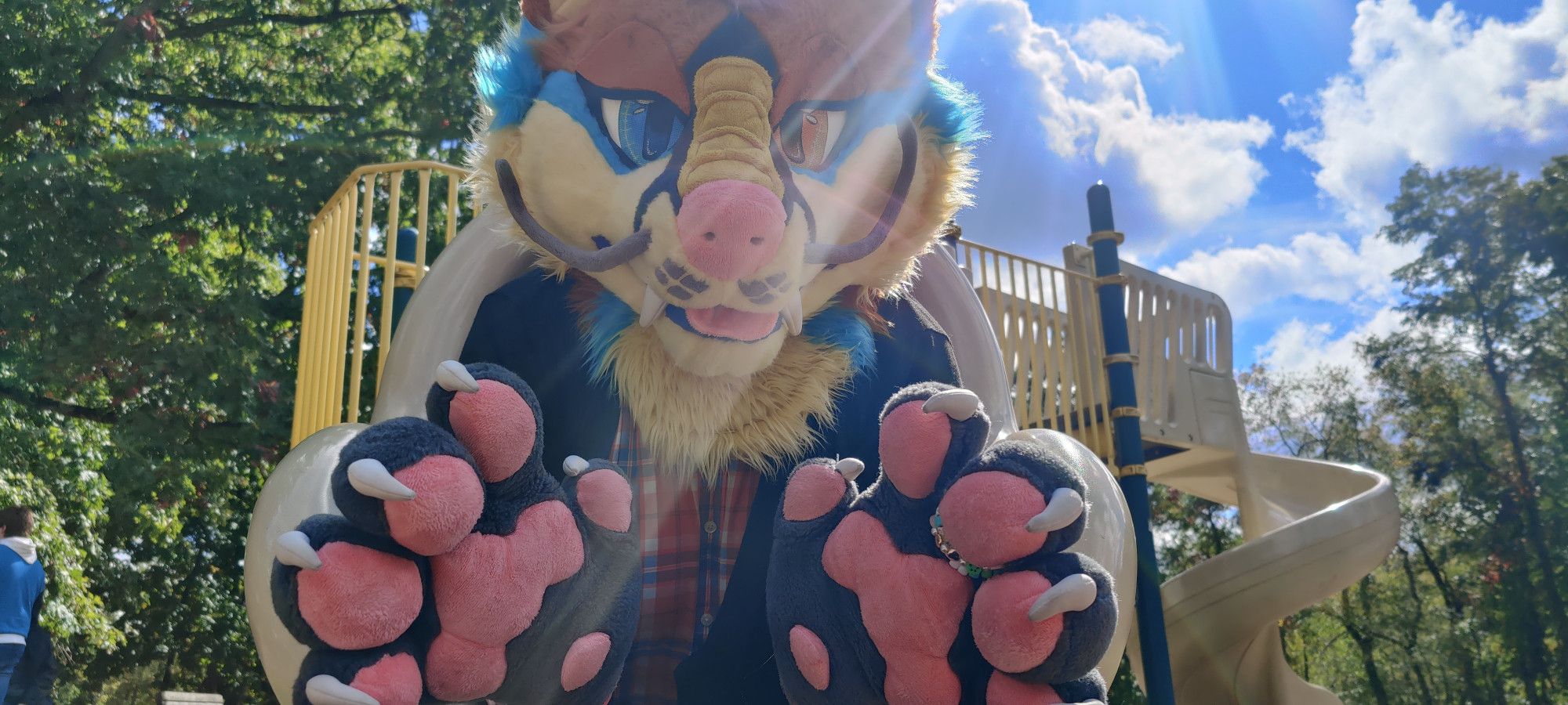 A dragon fox fursuiter emerging from the bottom of a slide with paws reaching out ahead. Photographed from below the subject fills most of the view but the rest of the playset is viable along with forest trees and a bright blue sky.