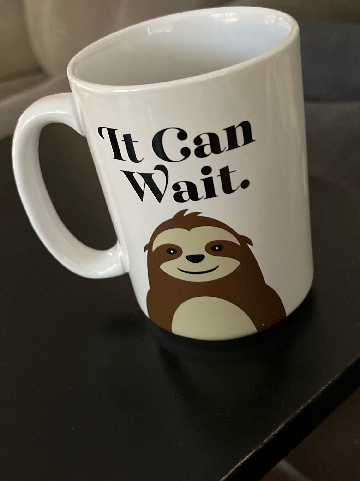 A white ceramic mug with the words, “it can wait” in black text. A graphic of a smiling sloth is below the words.