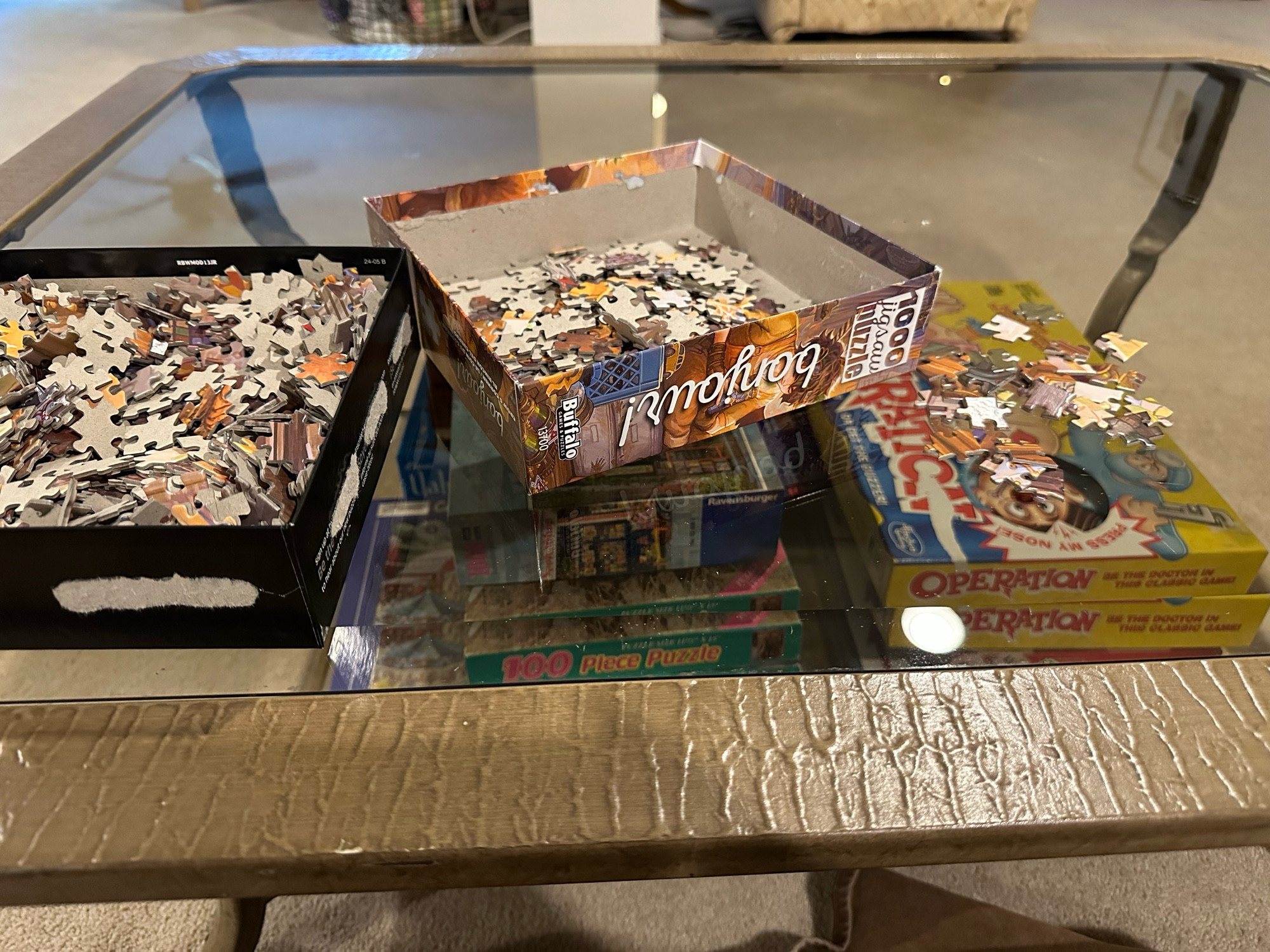 The image shows a glass coffee table with an opened jigsaw puzzle box. Pieces are spread out in both halves of the box, and some pieces are scattered on the table. Below the glass surface, there are board games like “Operation” and other puzzles visible. The table has a textured wooden frame, and the setting appears to be a living room.