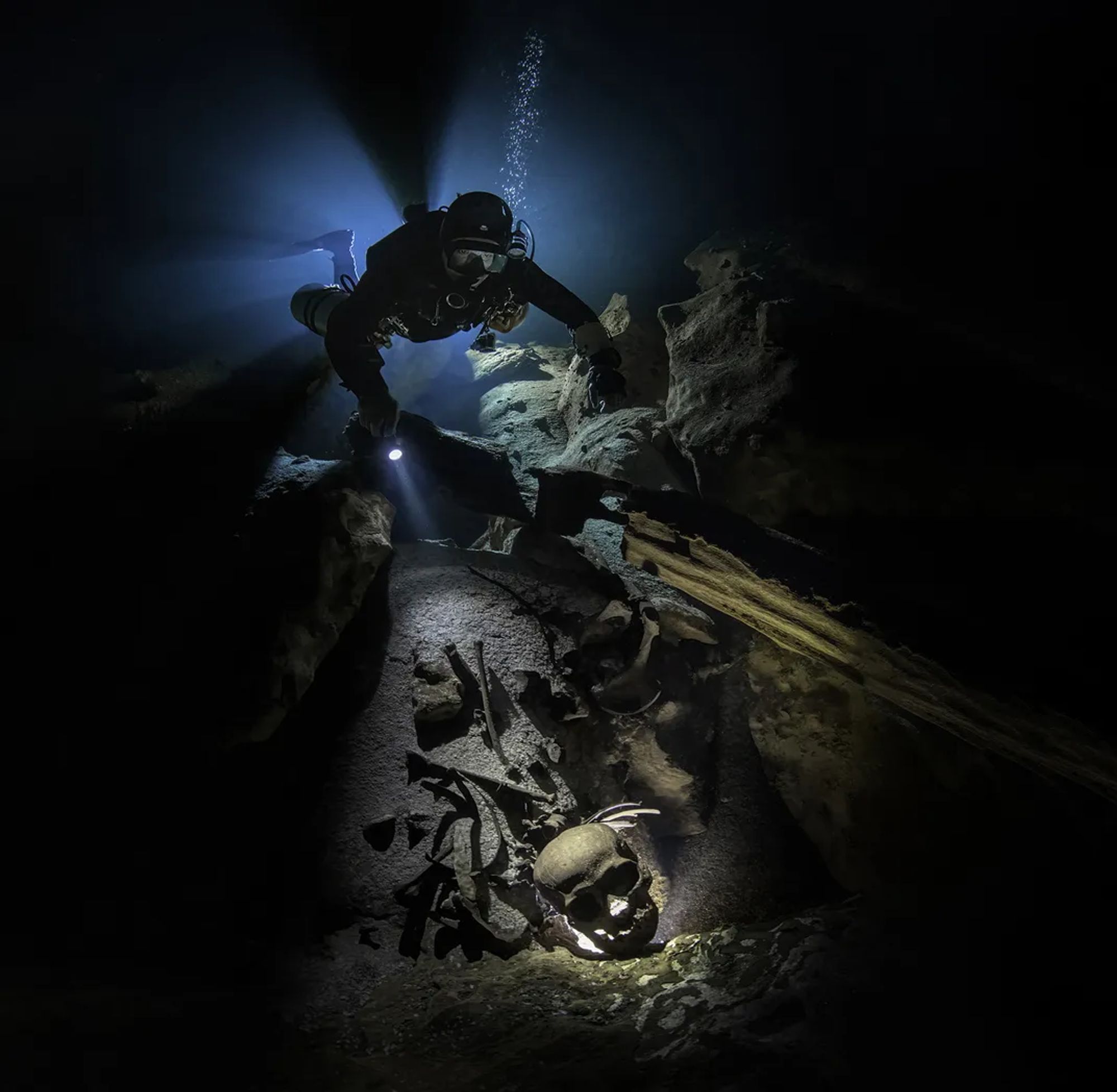 The image shows a diver illuminated by flashlights on their belt illuminating a mayan skull with the help of their flashlight.