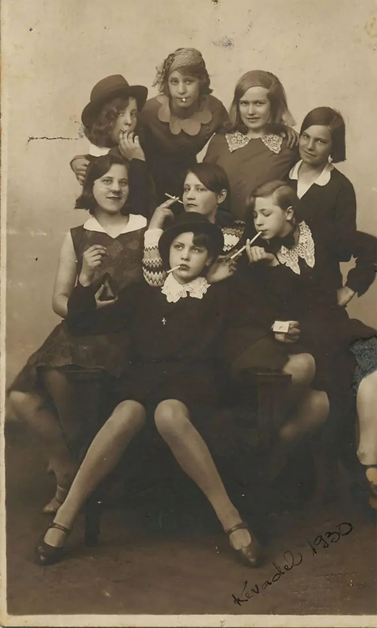 alt-text via GPT — "The image shows a group of eight girls from 1930 posing together. They are stylishly dressed, with some wearing hats and lace collars, and several are playfully pretending to smoke. Their expressions range from amused to contemplative, giving a lighthearted and candid feel."