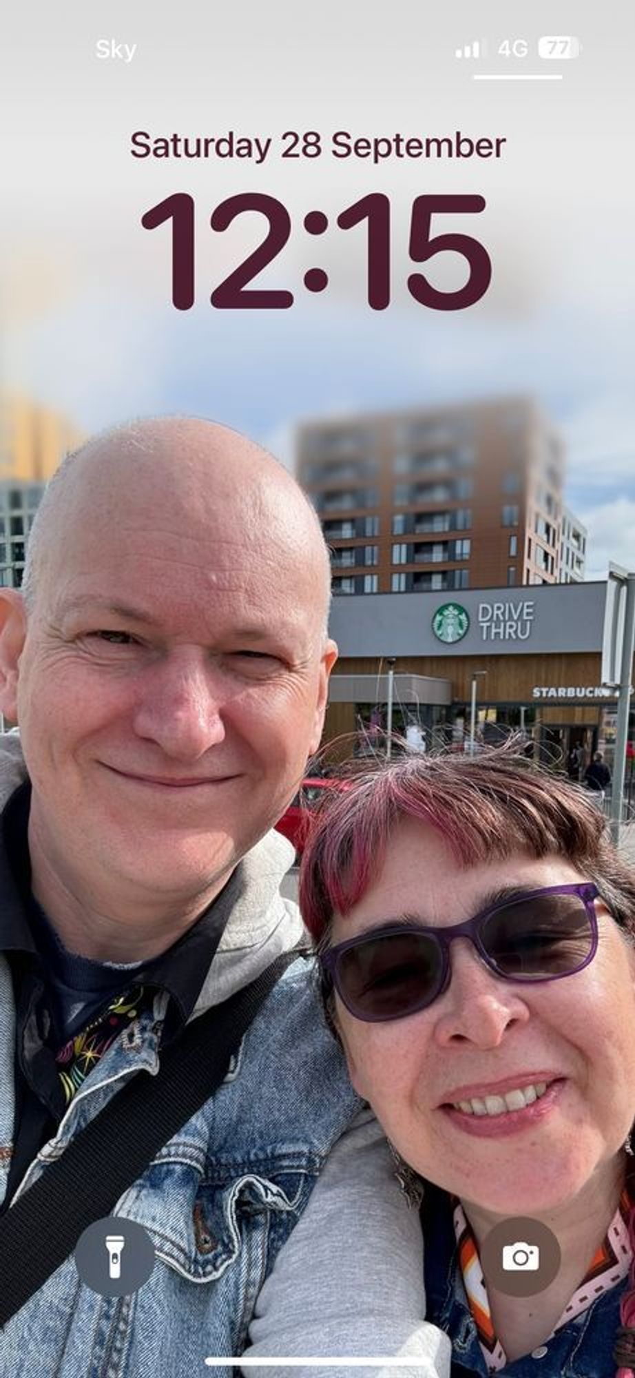 A man and a woman with glasses smile for the camera in a self portrait. Behind them is a drive through cafe and high rise buildings.