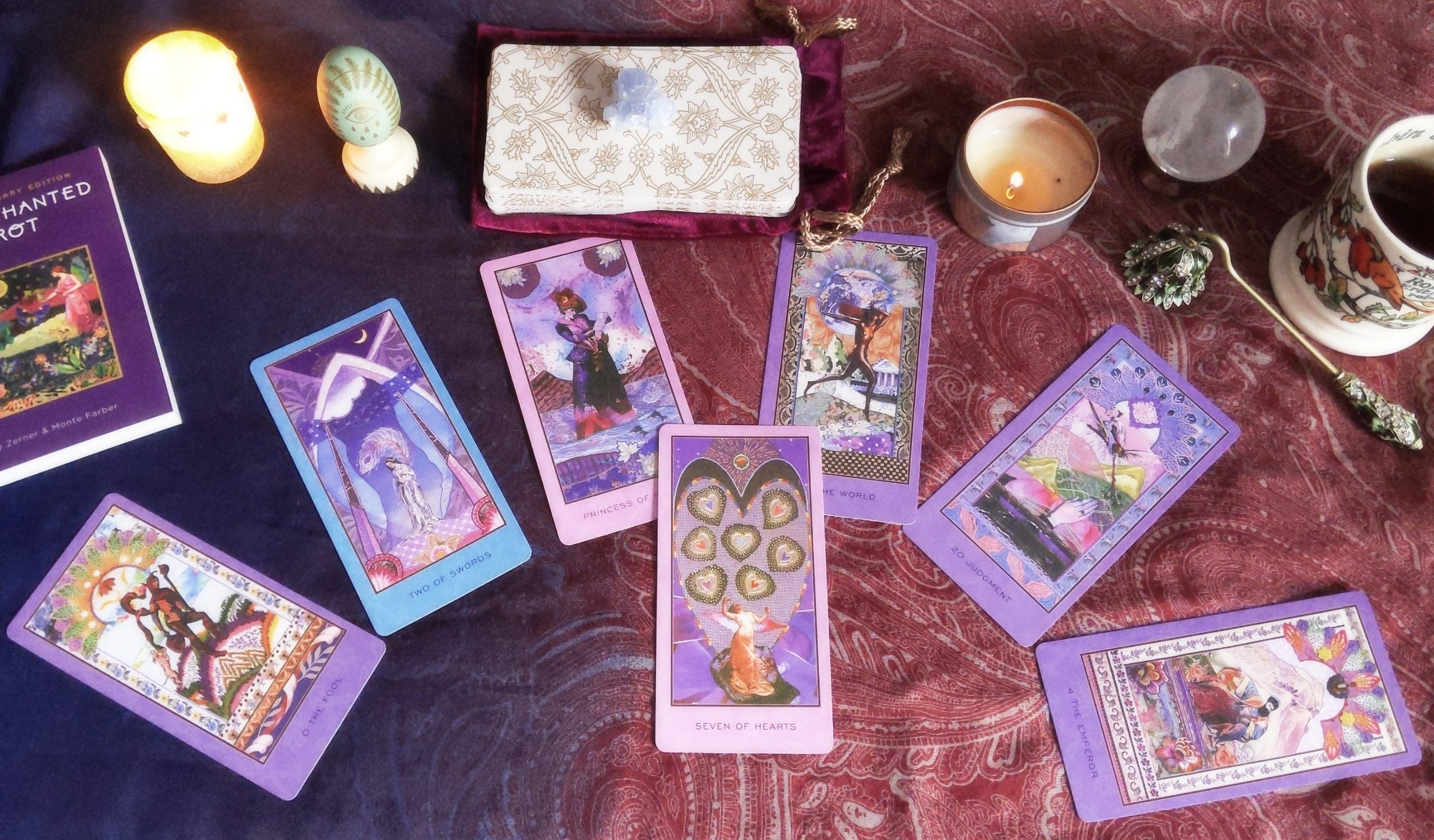 Photo of a Tarot spread using the Enchanted Tarot deck. Seven cards are laid out over a patterned scarf: The Fool, 2 of Swords, Princess of Hearts, The World, Judgement, The Emperor, and Seven of Hearts. The deck's guidebook, a couple of candles, a candle snuffer, a crystal ball and a mug of tea are also on display as decor.