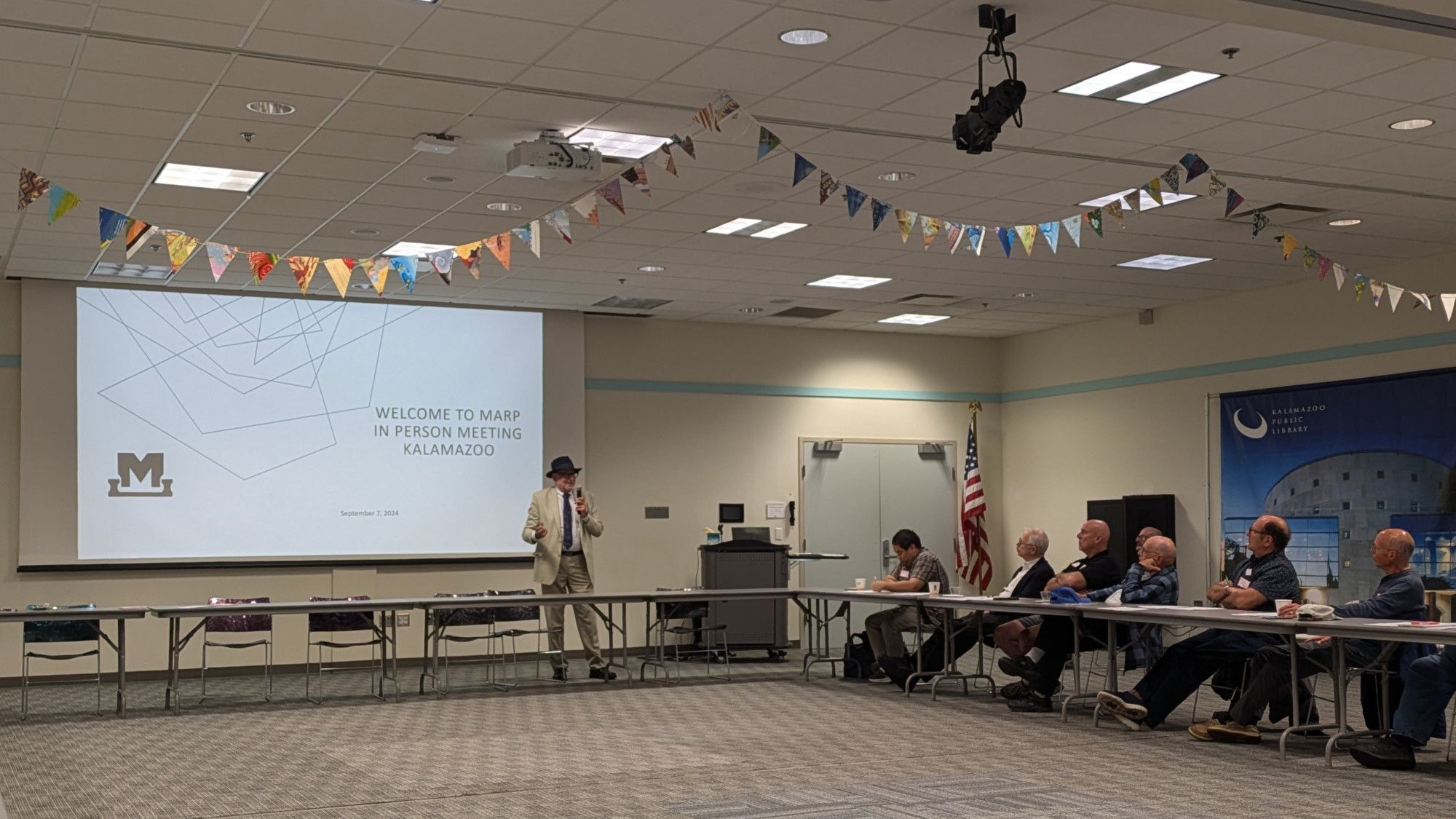 Mark Buckley, in a tan suit and black top hat, addresses meeting attendees in front of a projector slide reading "Welcome to MARP In-Person Meeting, Kalamazoo".