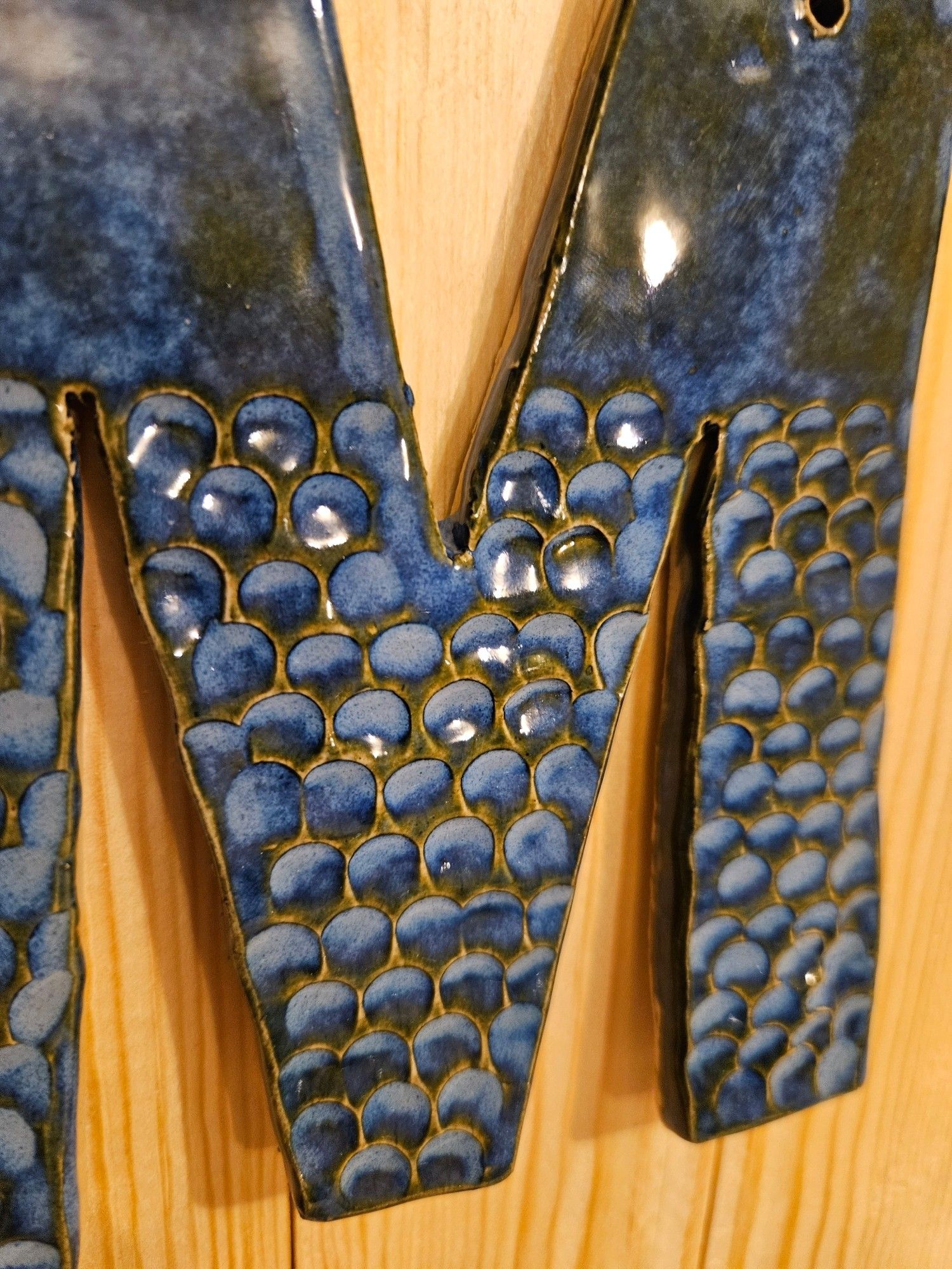 A ceramic letter M. It has a fish scale texture and is a bright deep blue that turns pale blue where it is thicker. There are spots that have a brown hue to it. It's a very organic color pattern.