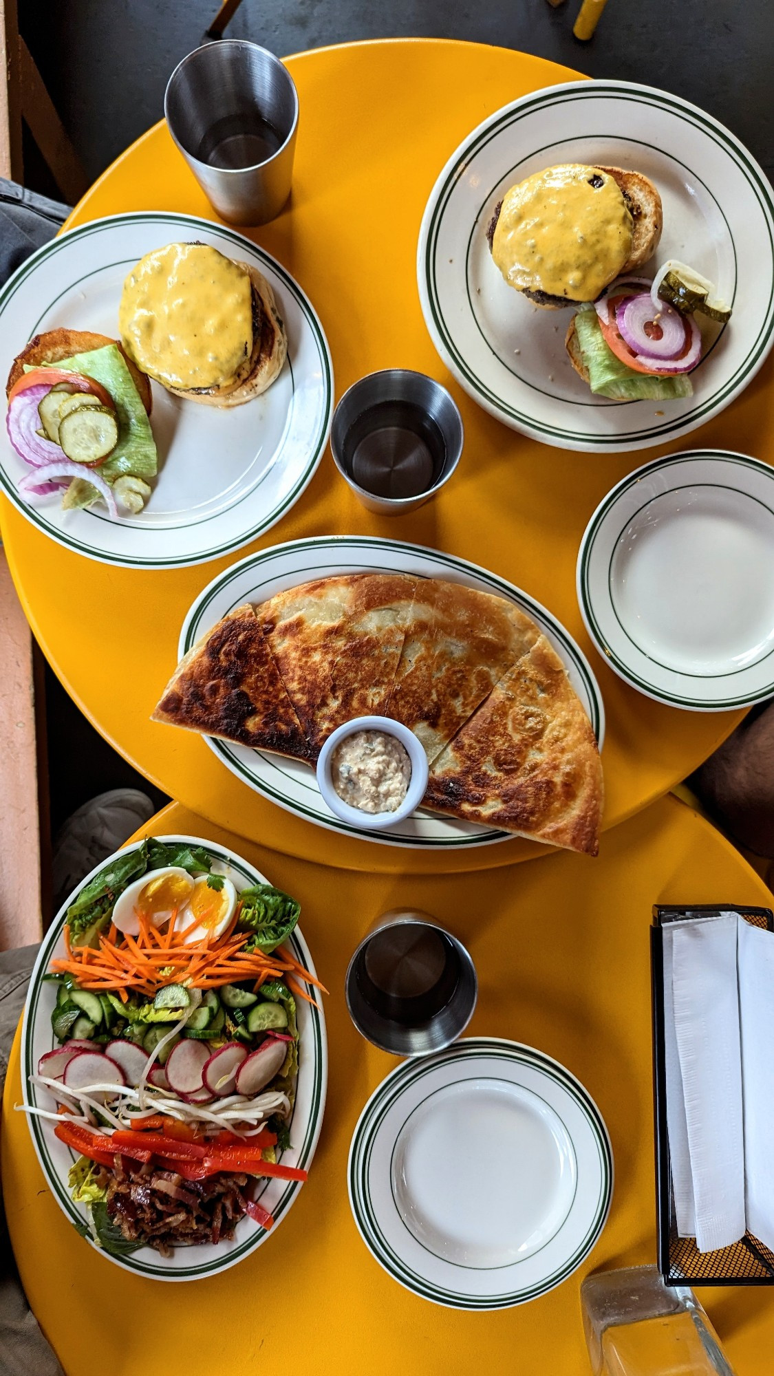 Top down image of beef cheeseburgers with lettuce, tomato, red onions, and pickles, a mushroom quesadilla, and a refreshing vinaigrette dressing Thai salad with pickled vegetables on yellow circular tables