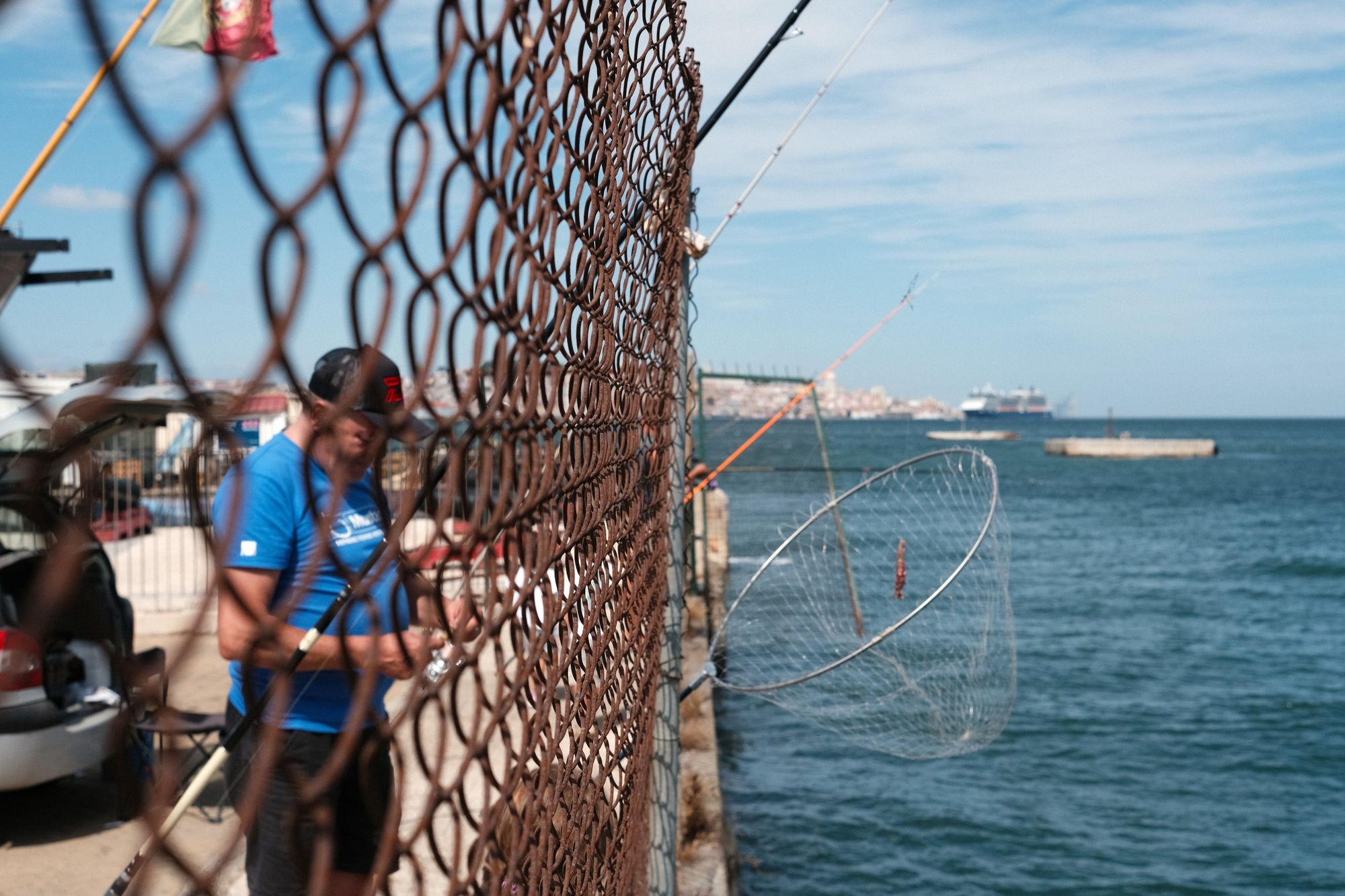 On the left, a fisherman preparing his fishing rod, seen through a chain link fence. to the right the Tagus river and the city of Lisbon in the background.