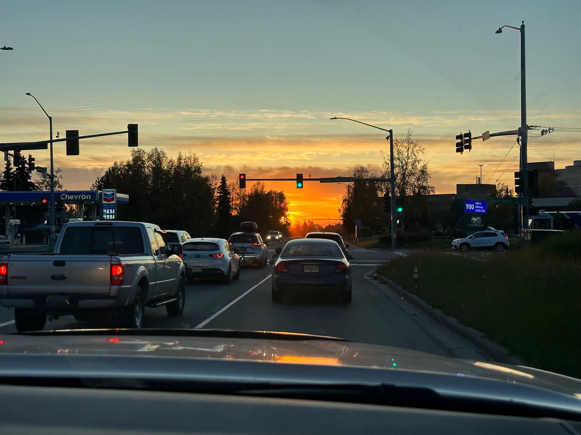 several cars are stopped at a four-way intersection stoplight. the photo is taken in the perspective of a passenger in the third car in line at the light, heading west towards the sun. the sky is colored a deep, molten orange close to the horizon. clouds slightly shroud the sun and are colored a rich, golden ocher. the orange of the sky melts into a faint, pale blue as it reaches further up the skyline.