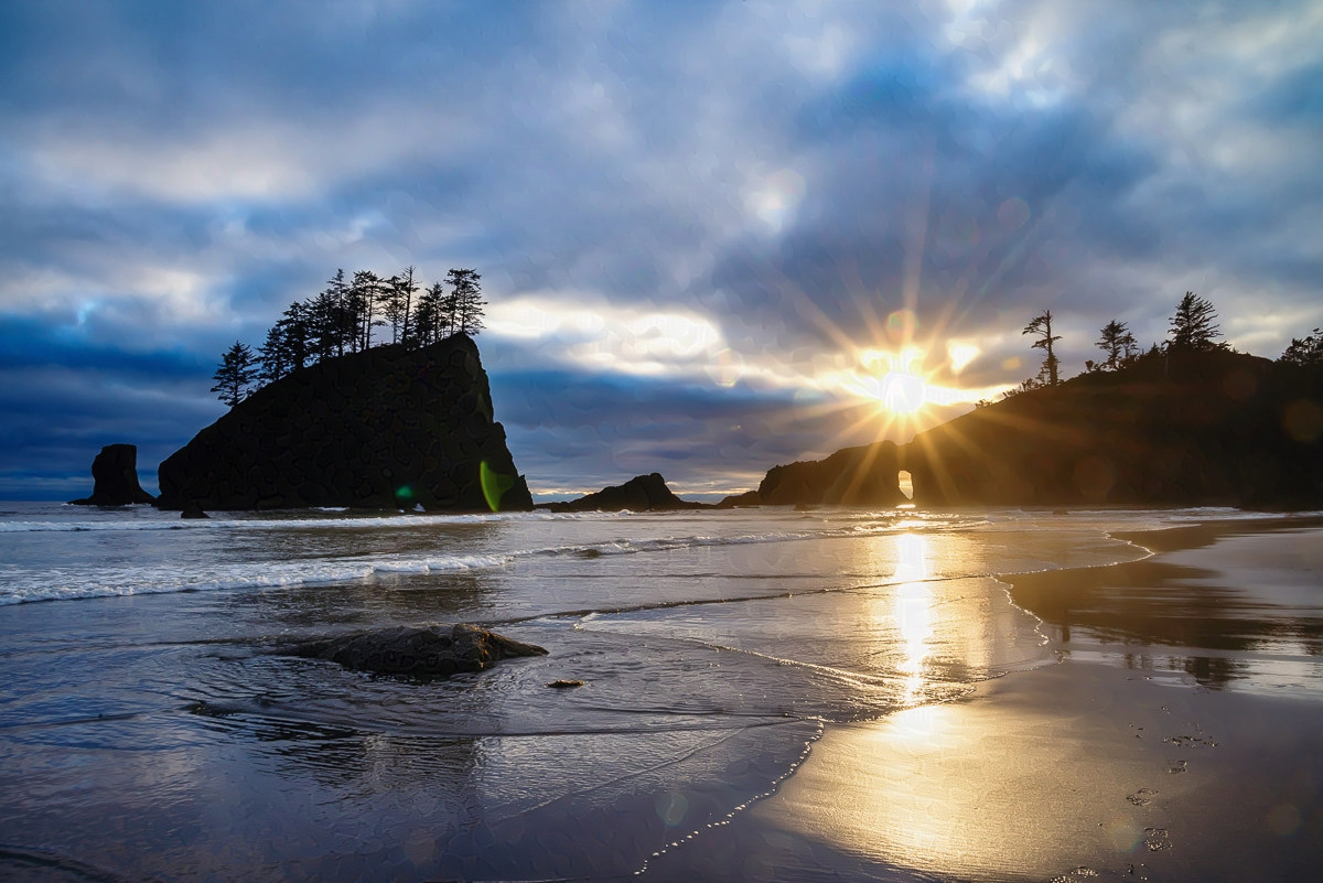 A sun-flare on a beach.

In the foreground, ocean waters come up gently on a sandy beach. The waves are tiny, just a couple of inches high, and the sand is wet from the tide coming in.

In the middle ground are some tall sea stacks. There are trees growing on top of them. There is a small arch in the sea stack to the right, and you can see through it.

The sun is just above the sea sick to the right and it has sun rays.

The sky is cloudy with heavy clouds. You can see a few scraps of blue, but it also looks like it might rain.