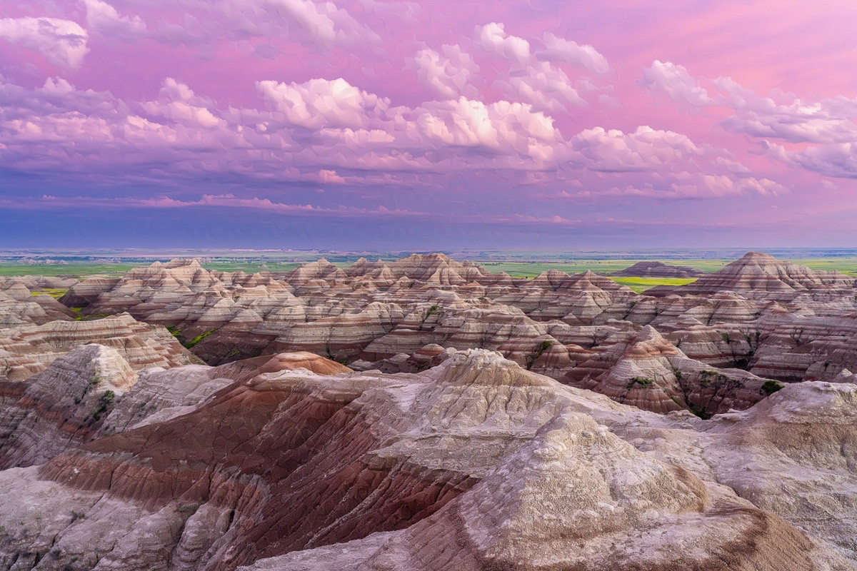 A view from an overlook. 

In the foreground, striped, low hills stretch into the distance. These are predominately brown, and the tops are lighter. Striations are on every hill, making them appear striped. 

In the far distance you can see the green and yellow of summer grasses and flowers.

The sky is pink with some white fluffy clouds. Toward the horizon the sky blends into more purple tones.