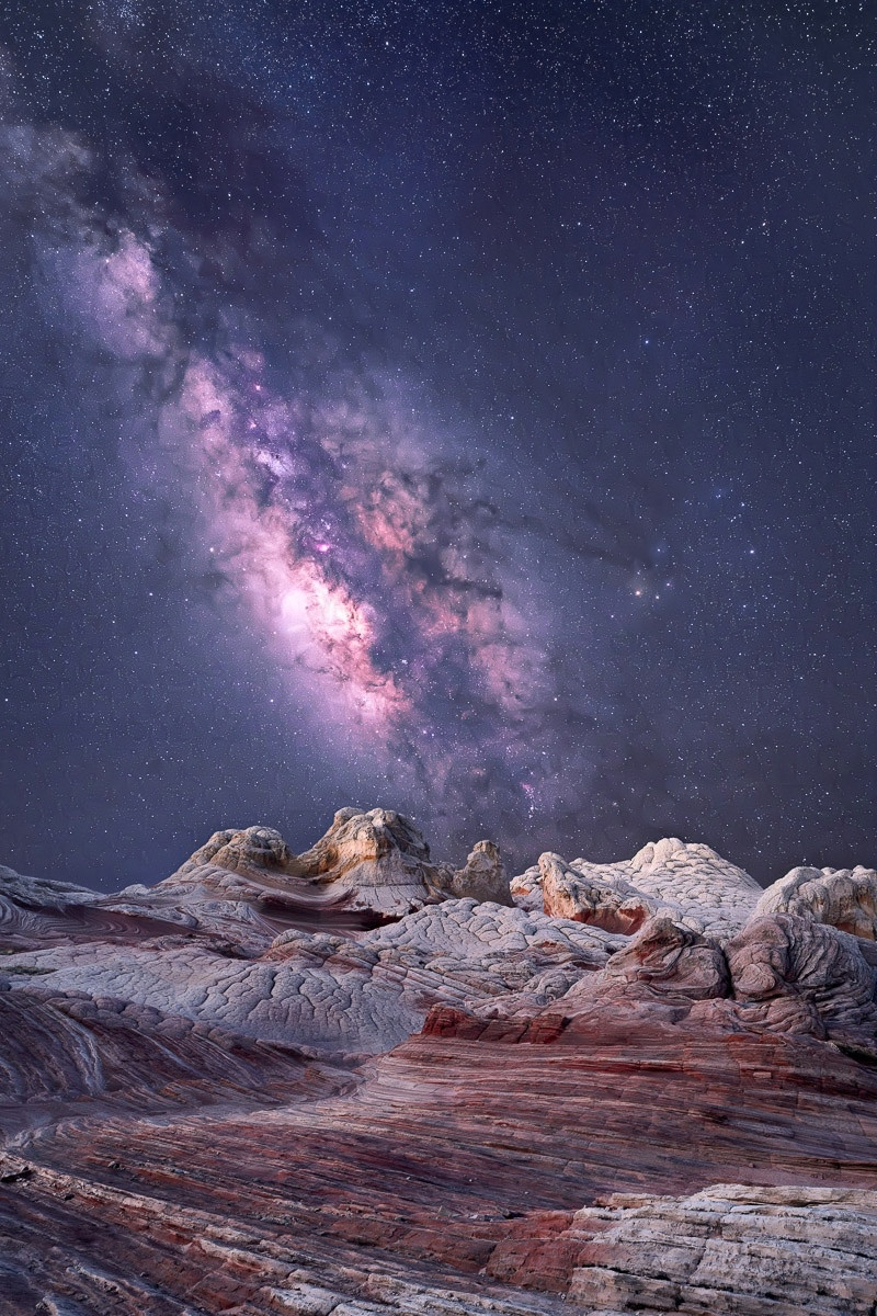 The Milky Way over the rocks of White Pocket.

In the foreground are twisted formations of rocks, with lines, grooves and protrusions everywhere. The rocks are white with red accents. In general, the rocks lead from the front to the back.

The Milky Way takes up the sky, gently arcing in from the upper left to the middle.

The photograph has deep purple hues.