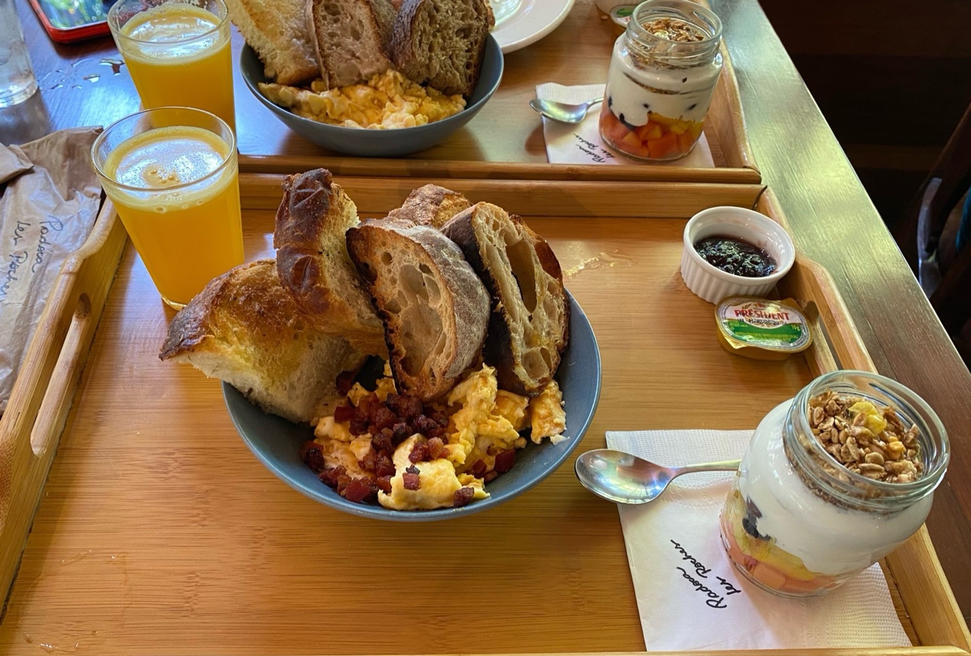 Foto de uma bandeja com brunch. Tem um copo de suco de laranja, um mini ramequim com geleia de frutas, uma mini manteiga de hotel, um pote de vidro com Kefir frutas e granola, e uma cumbuca com ovos mexidos, bacon e algumas fatias de pão