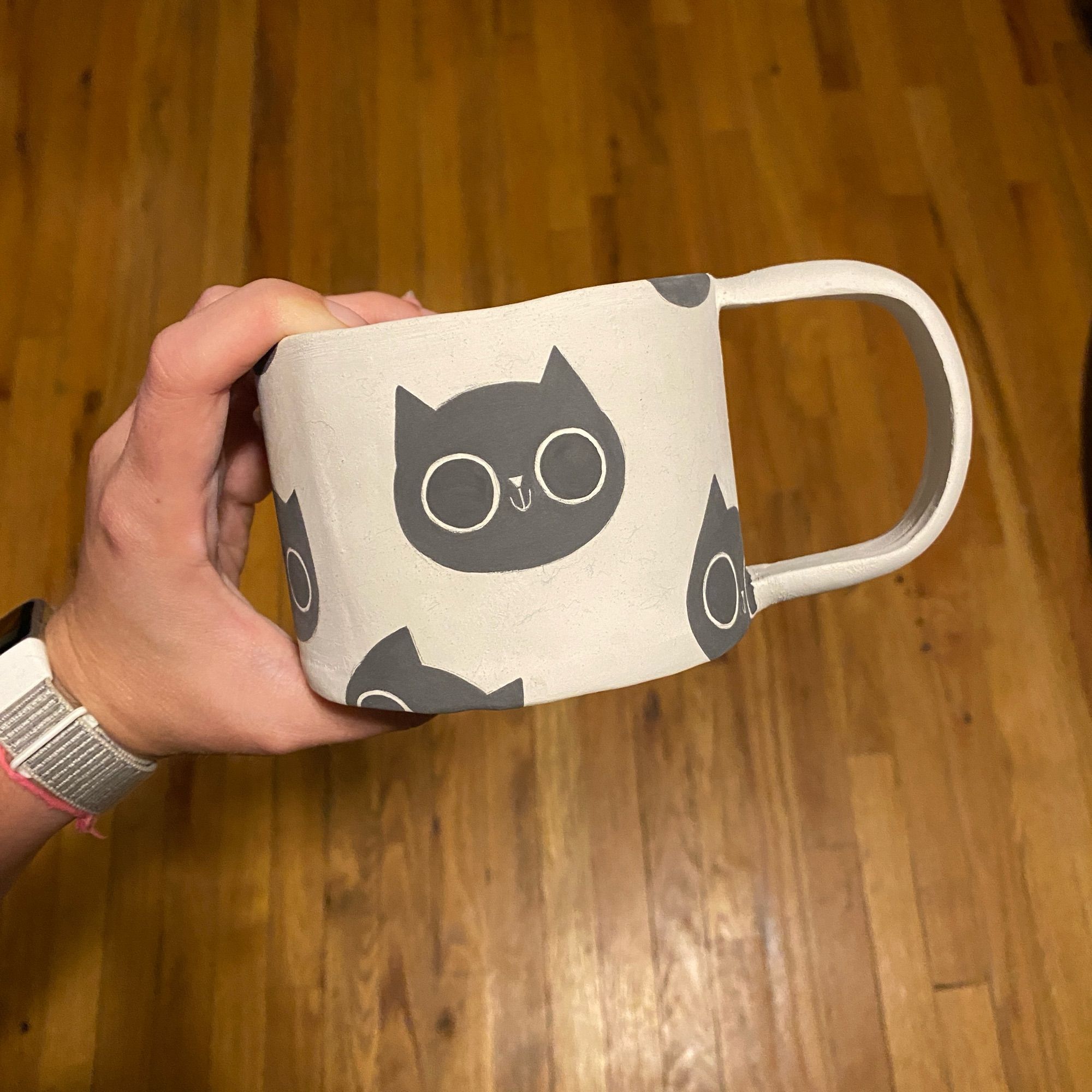 Hand holding a greenware ceramic mug with black cats all over the surface