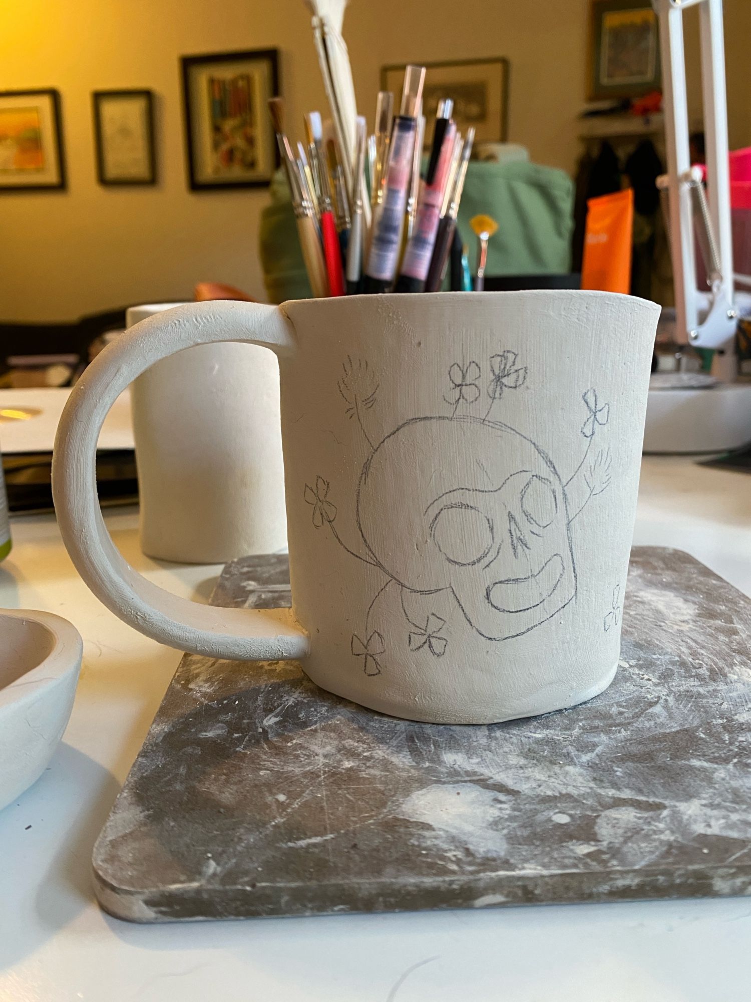 An unfinished ceramic mug rests on a wooden board with other unfinished pieces and brushes around it in the background