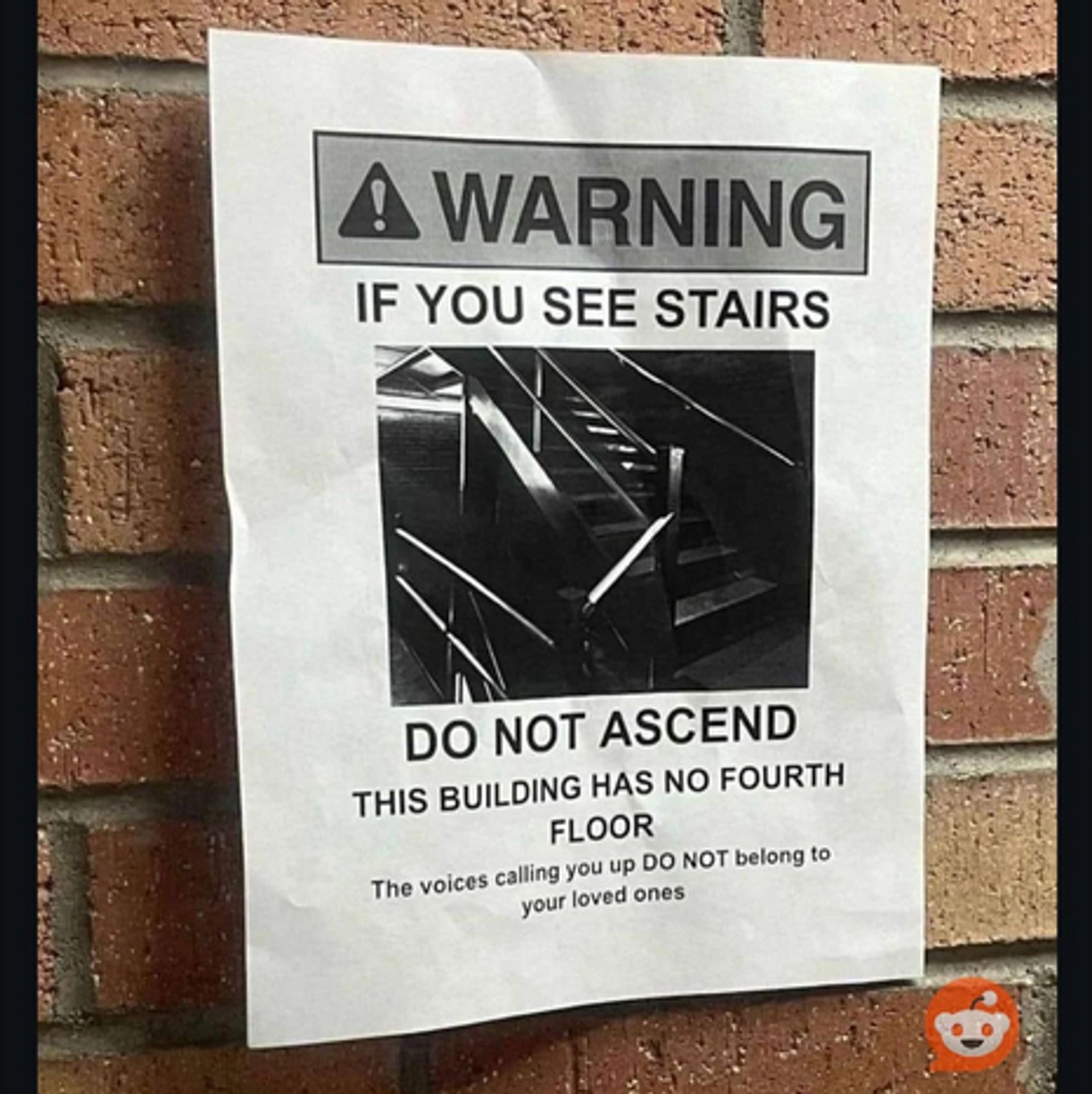 A black and white poster on a red brick wall. 
WARNING 
If you see stairs 
[photo of a dark stairwell] 
do not ascend. 
This buildling has no fourth floor. 
The voices calling you up DO NOT belong to your loved ones.