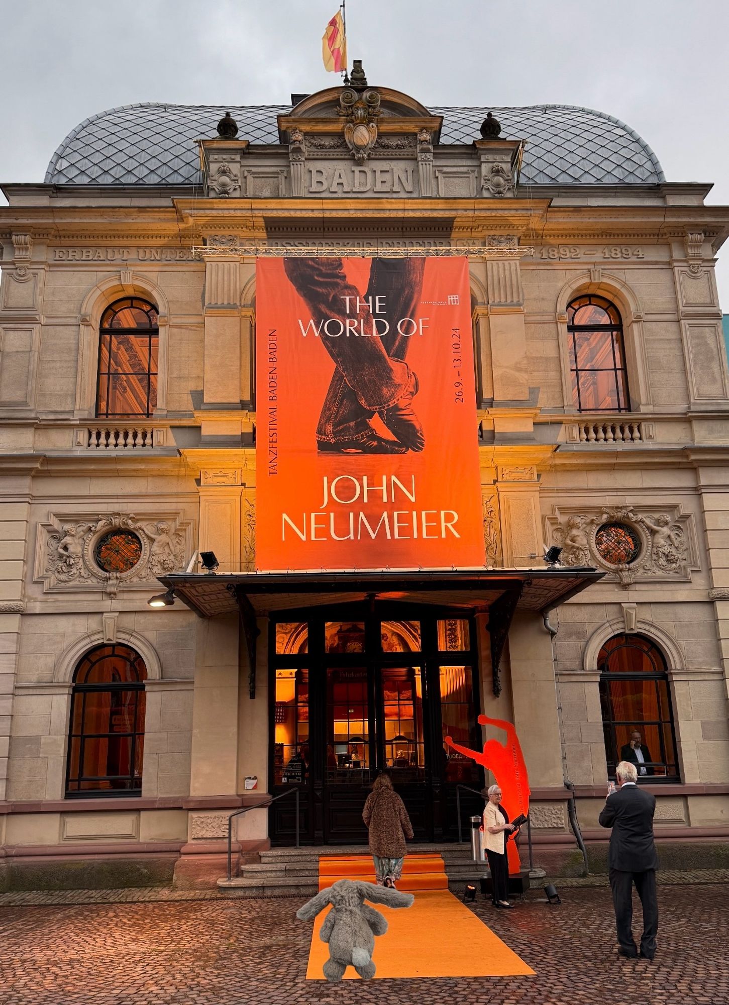 Festspielhaus BadenBaden mit Plakat "The world of John Neumeier oberhalb des Eingangs. Davor ein. orangefarbener Teppich mit reinmontiertem Hasi