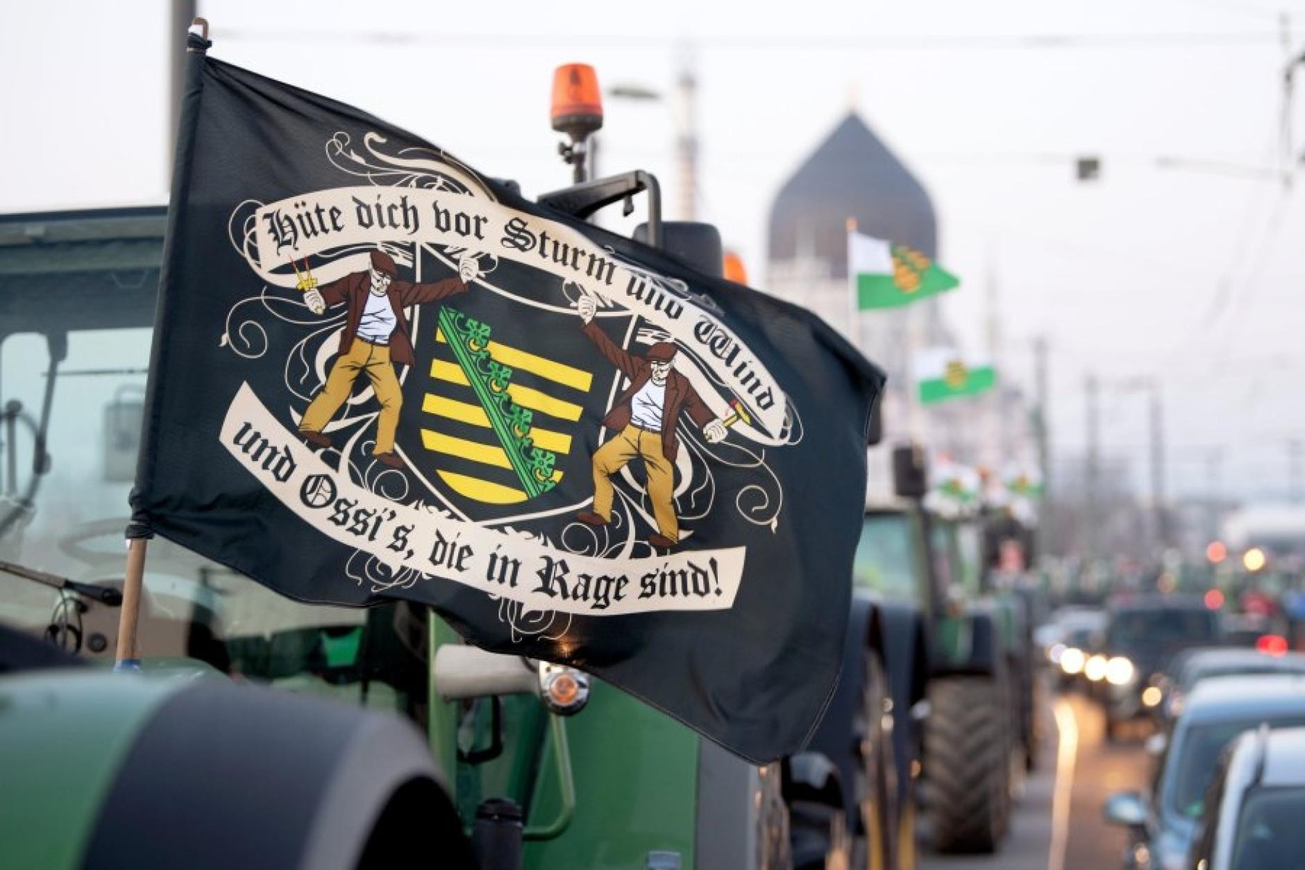 Foto aus Dresden, Straße mit Traktoren, im Vordergrund Traktor mit Fahne, Sachsenlogo, Aufschrift "Hüte dich vor Sturm und Wind und Ossis die in Rage sind!" In Fraktur.