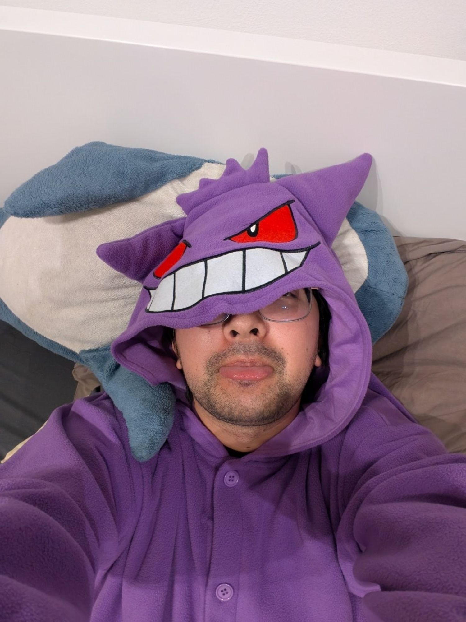 Me, in bed, wearing a Gengar onesie. Gengar's grin is covering the upper part of my face. I've stuck my tongue out slightly.