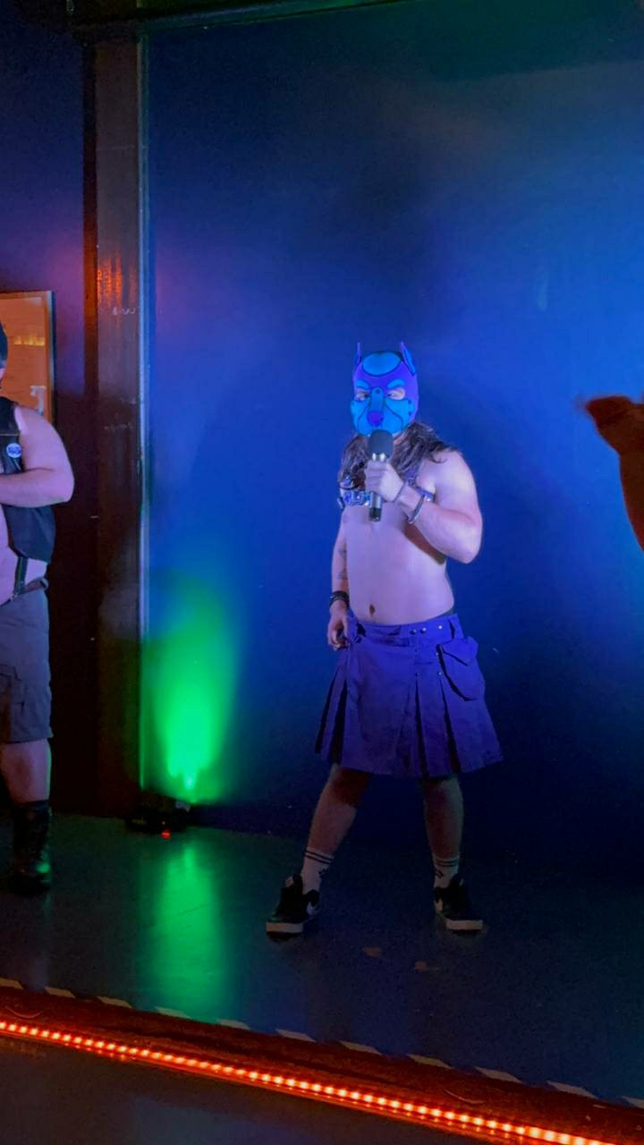 Leather boy and human pup singing on stage. Leather pup is wearing a purple kilt and a purple leather harness with a blue and purple hood