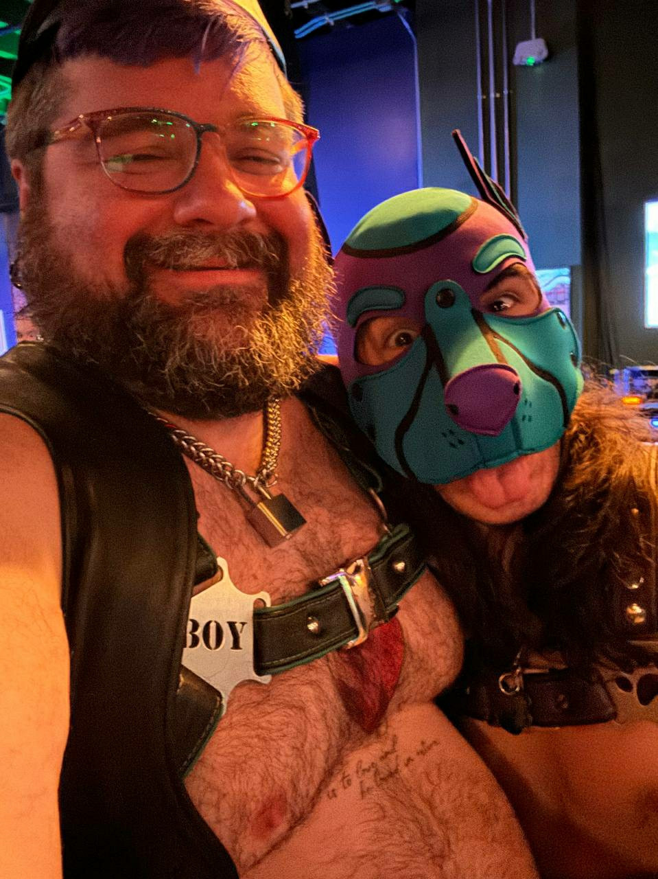 Picture of two cuties: Left is a bearded man in glasses with leather harness that says "boy" and is wearing a chain collar with a lock on it. Right is a pup in a purple and blue hood with their tongue sticking out and a purple harness with paw prints on it