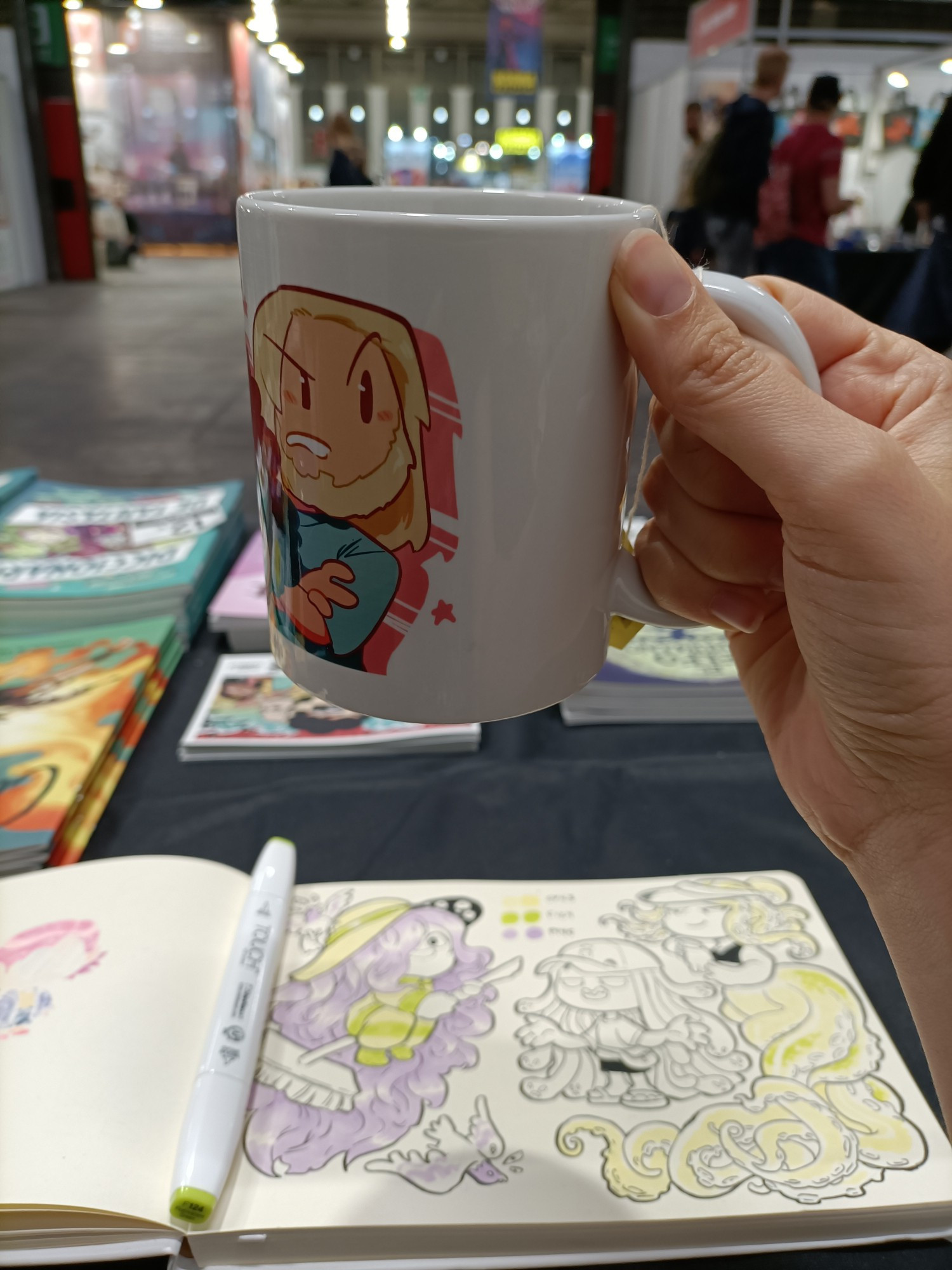 Foto hecha desde dentro de un stand de una mano sosteniendo una taza de té, de fondo borroso se ve un cuaderno con dibujos y gente paseando