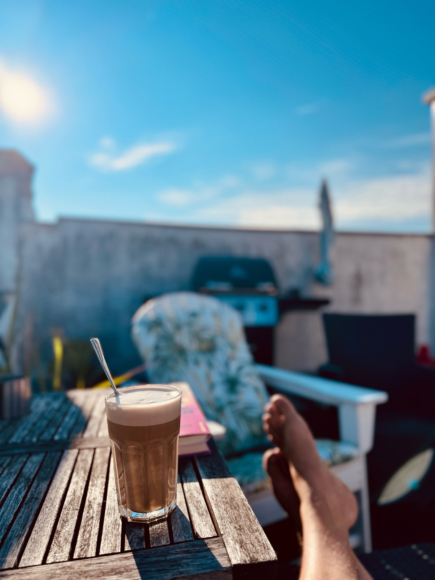 Ein Latte Macchiato in einem klaren Glas steht auf einem Holztisch im Freien. Im Hintergrund sind ein Buch, ein Stuhl und eine Grillstation zu sehen. Eine Person entspannt mit hochgelegten Füssen in einem Liegestuhl. Der Himmel ist klar und sonnig.