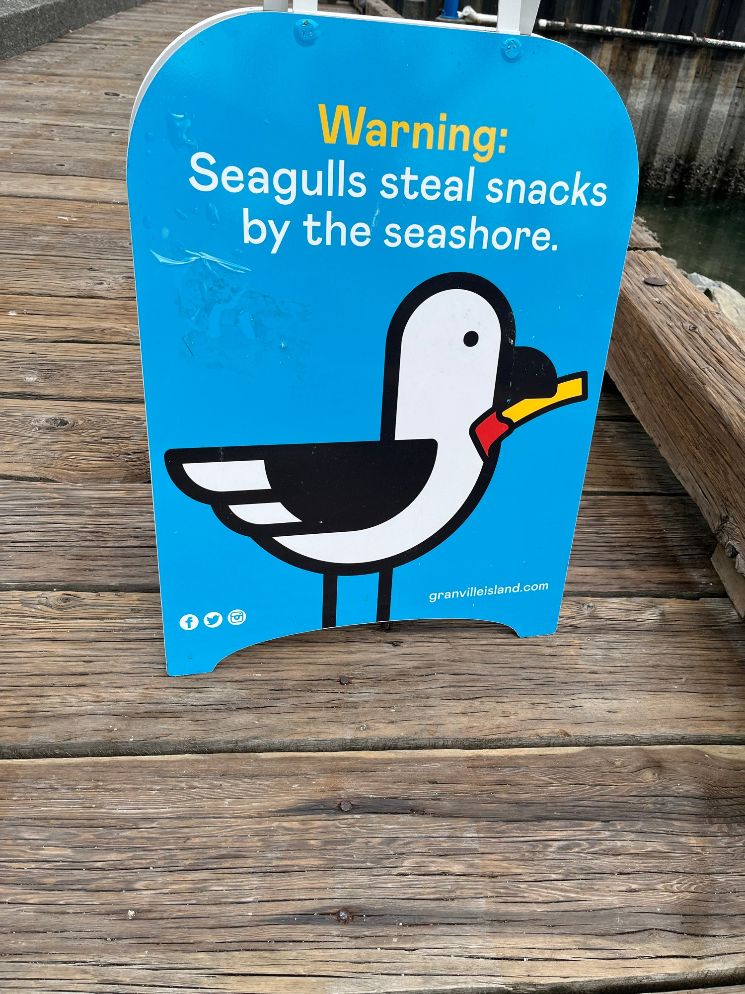 Blue poster standing on wooden boardwalk, oceanside, outside Granville Island market. Image: cartoon white seagull, yellow french fry tipped w red ketchup in its beak. Text: “Warning: Seagulls steal snacks by the sea shore.”