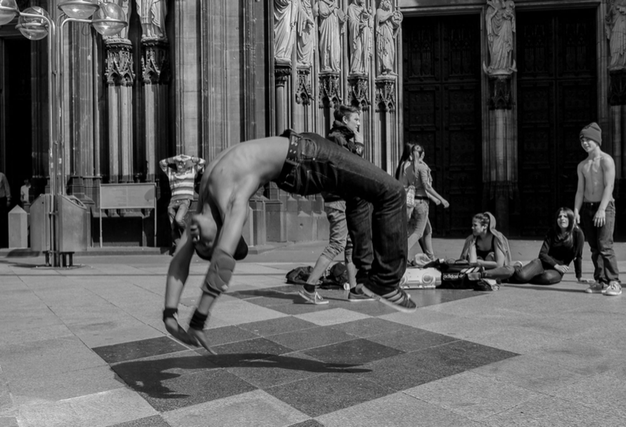 Ein Schwarz-Weißbild im Querformat. Ein Jugendlicher macht Breakdance auf der Kölner Dom Platte und ist gerade in der Luft weil er rückwärts von den Füßen auf die Hände springt. Im Hintergrund sind weitere Jugendliche und der Dom.