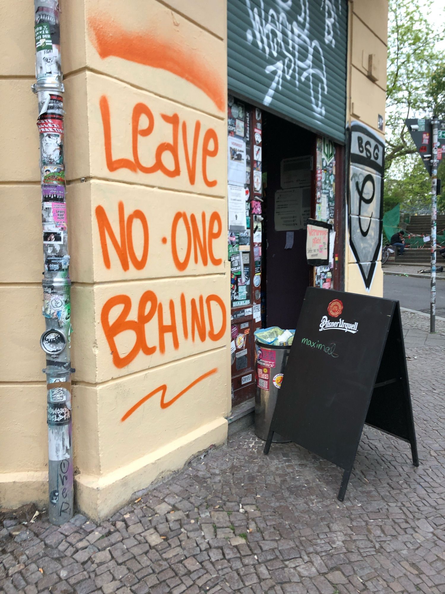 An einer Wand steht groß "LEAVE NO ONE BEHIND" an einem Laden. Davor steht ein aufstellen des Ladens / spätis und Teil der Straße sind zu sehen.