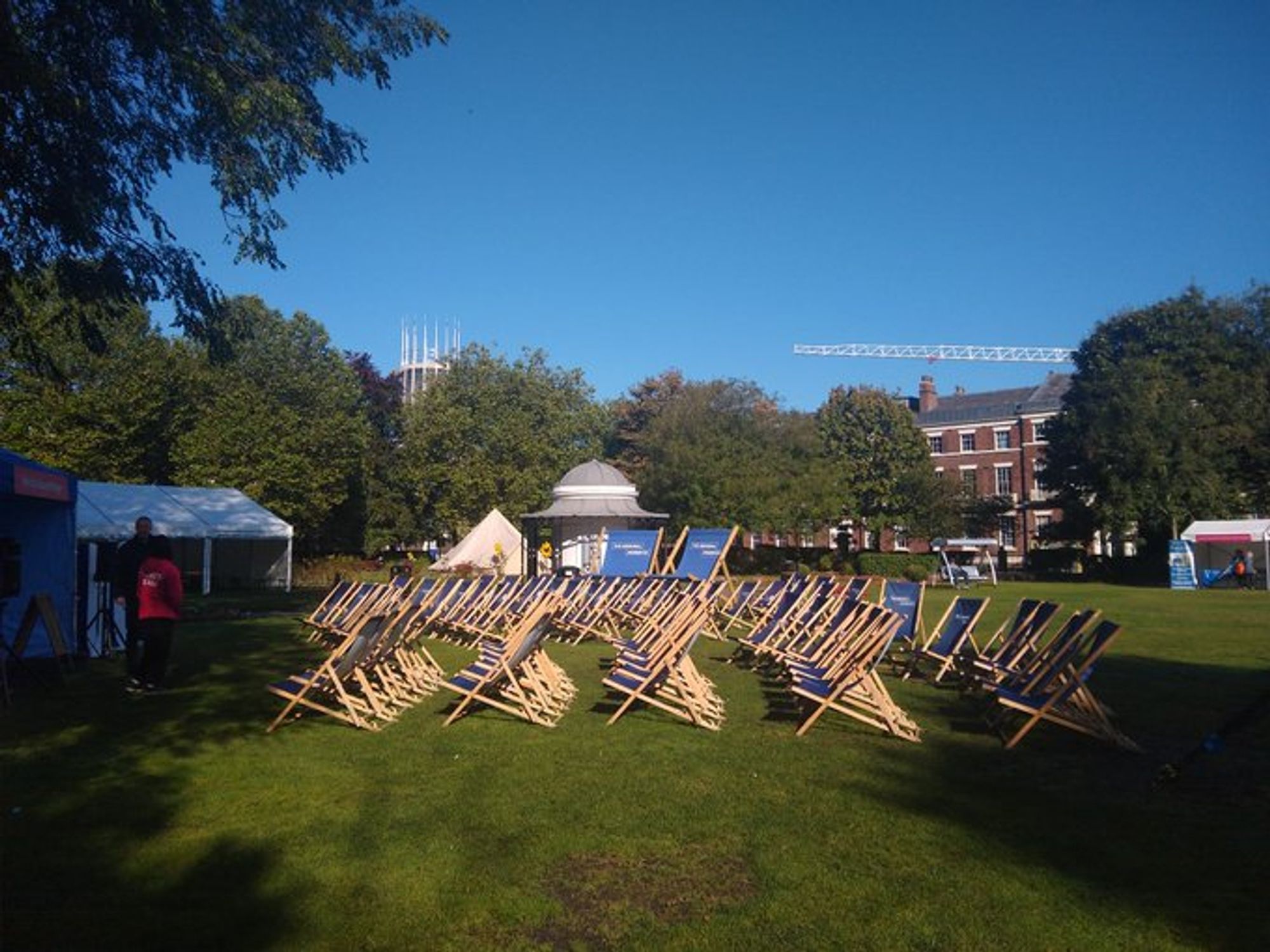 Rows of blue sun loungers in the sunshine on the lawn of Abercromby Square. The original gazebo as well as marquees for Welcome Week are in the background. Further back are the trees surrounding the square with the Catholic Cathedral peeking out over the top in front of a blue sky.