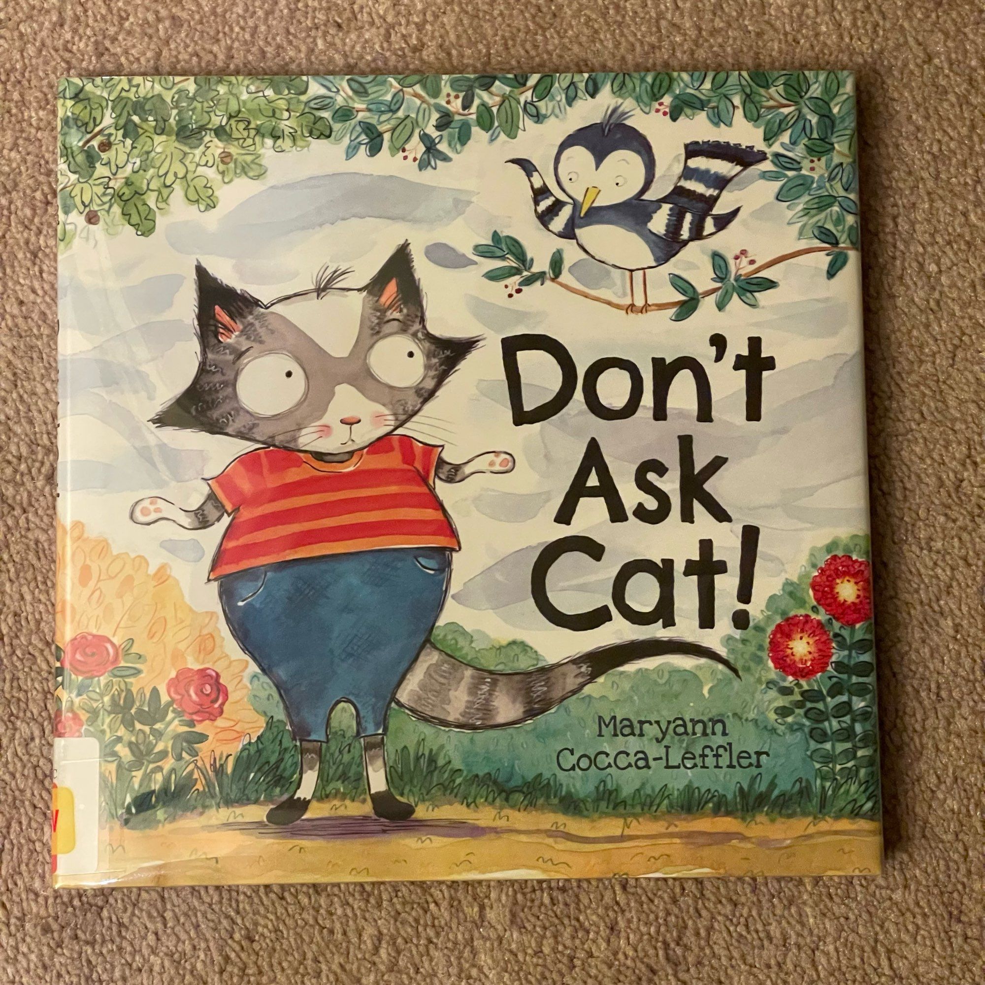 Photo of the front of picture book “Don’t Ask Cat!” that is laying on a tan carpet. The cover depicts an outdoor path with sky, tree branches, grass, bushes, flowers, and a dirt path. A cat standing on back legs and wearing a striped shirt and blue pants is shrugging and looking at a shrugging blue and white bird who is perched on a tree branch.