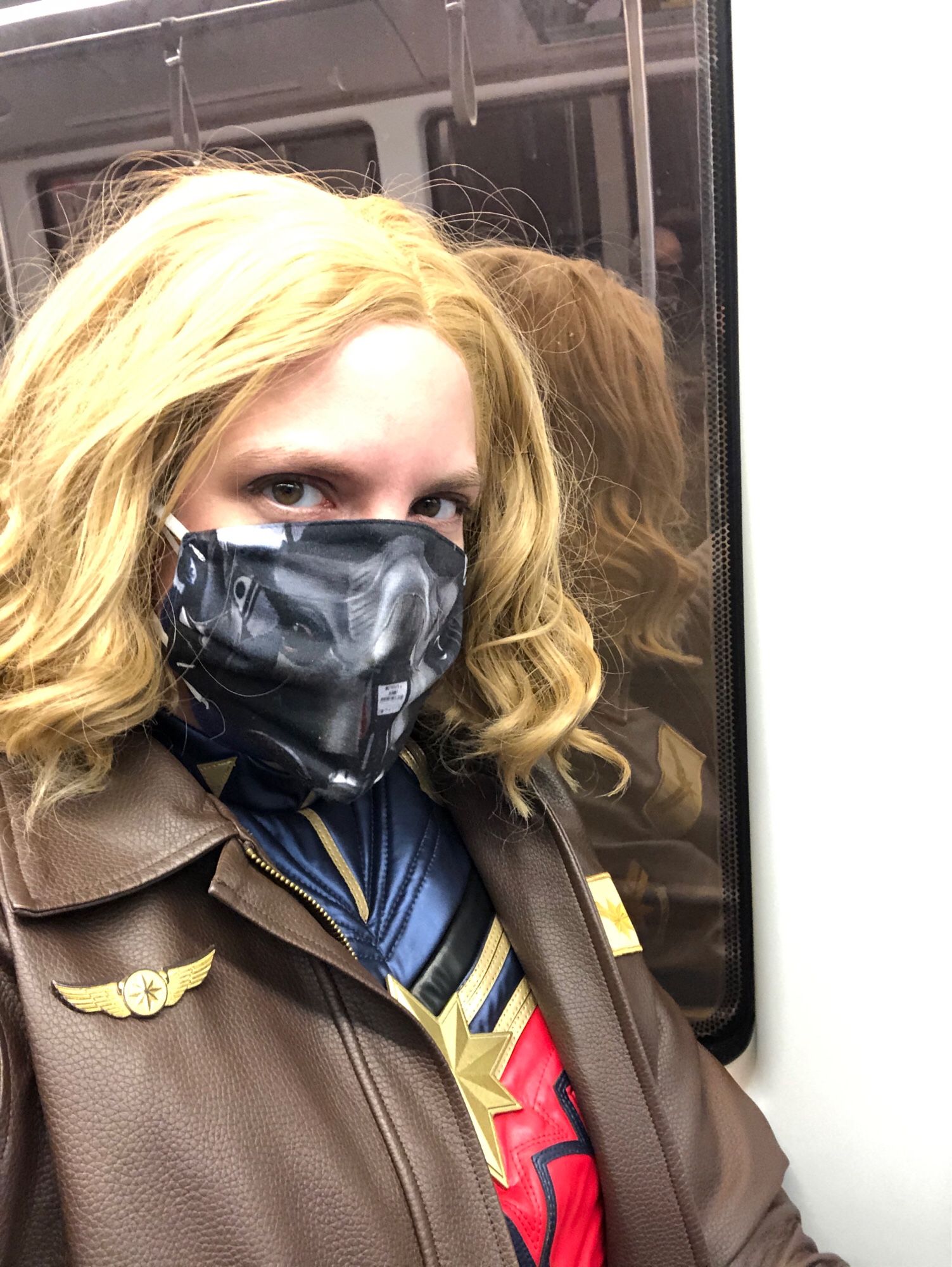 Captain Marvel in her suit and bomber jacket, riding on a light rail train. Her face mask depicts the O2 mask worn by fighter jet pilots.