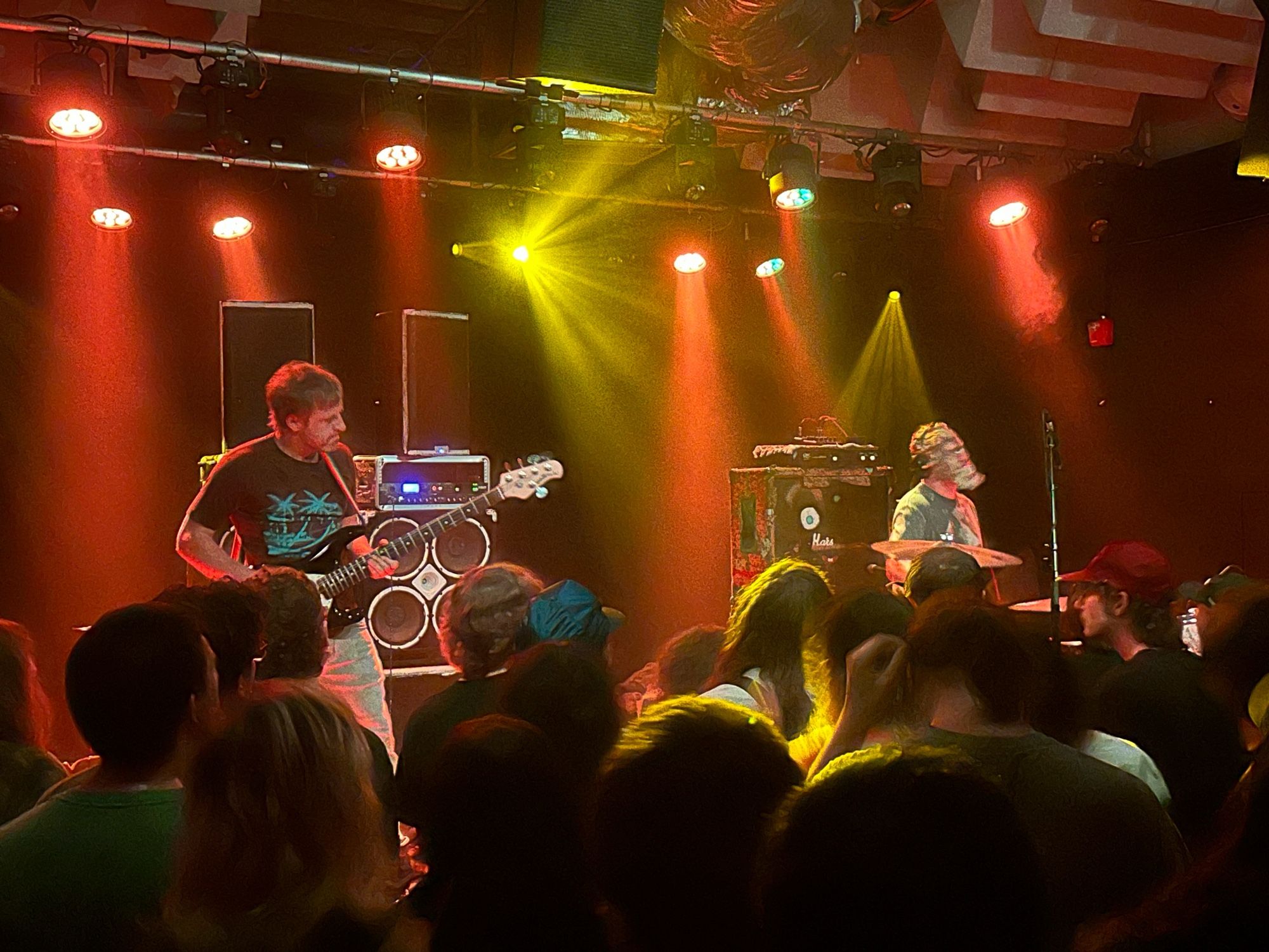 A man playing bass while another man wearing a mask is on the drums. The crowd is standing in front of them in movement. The stage is bathed in orange and yellow light.