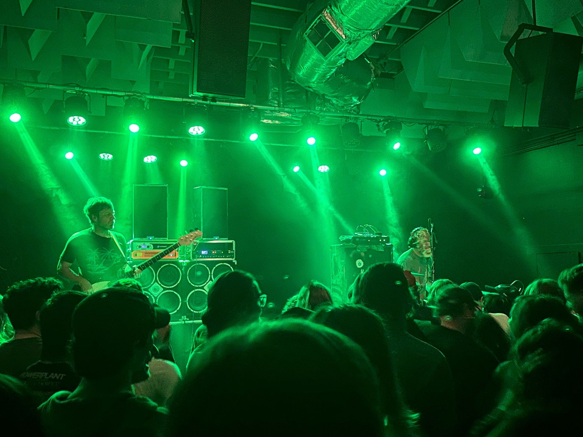 A man playing bass while another man wearing a mask is playing the drums. The back of the crowds head is visible in front. Everything is green from the stage lights.